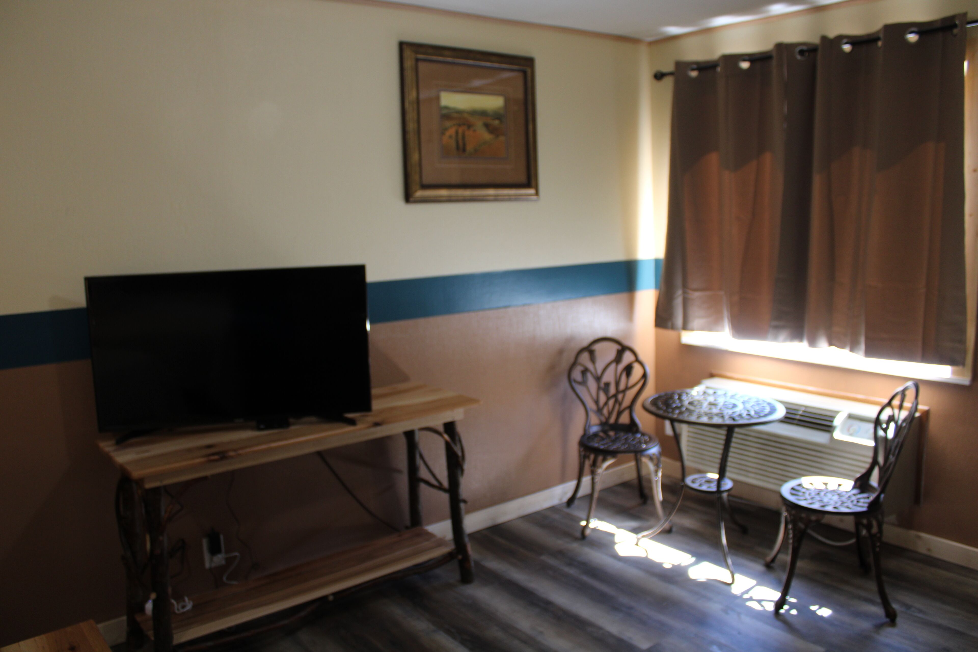 A room with a television on a wooden stand, a framed picture on the wall, a window with brown curtains, and two chairs around a small table.