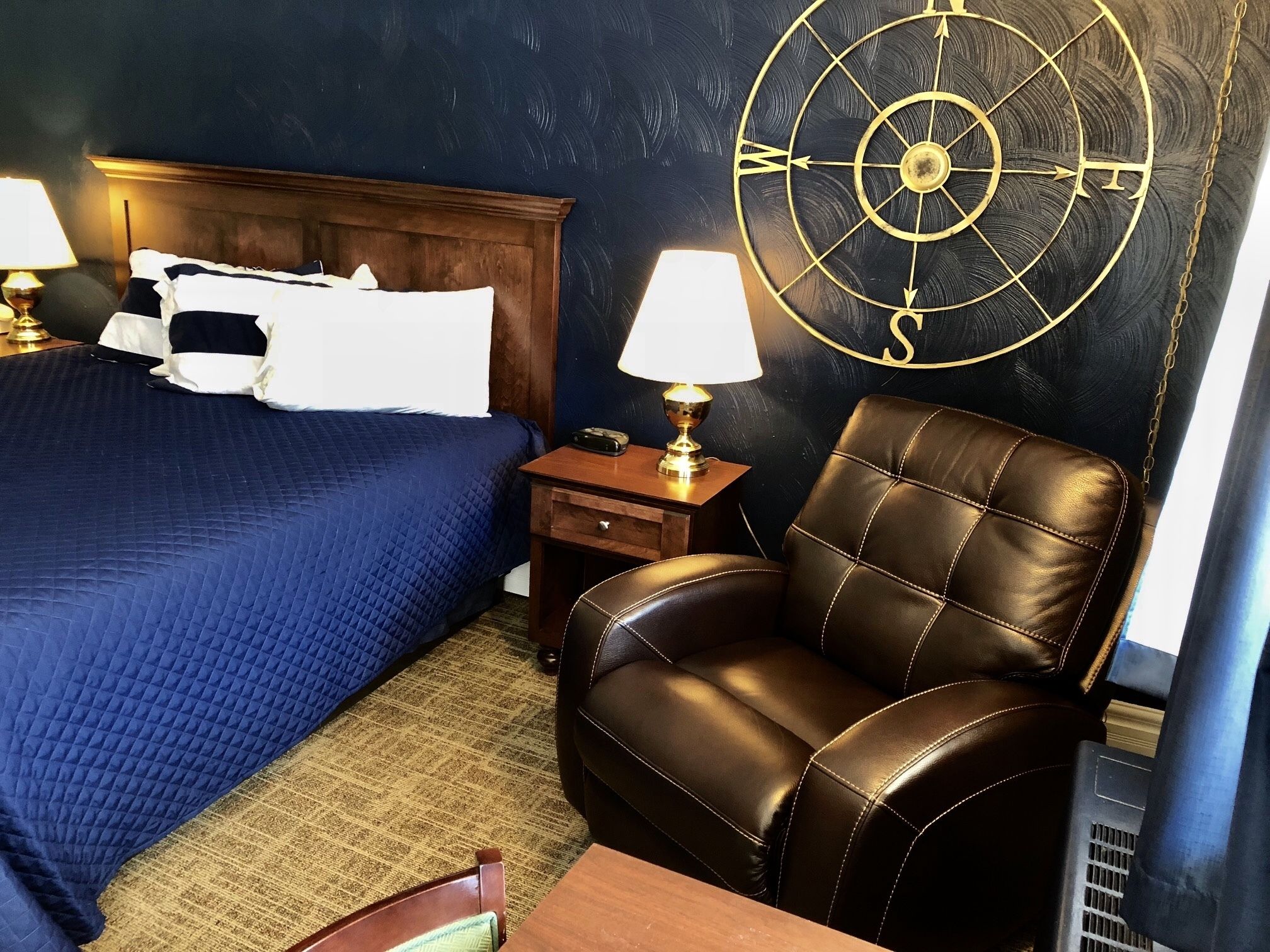 Una camera d'albergo con un letto, una poltrona in pelle, un comodino con una lampada e una sveglia a parete.