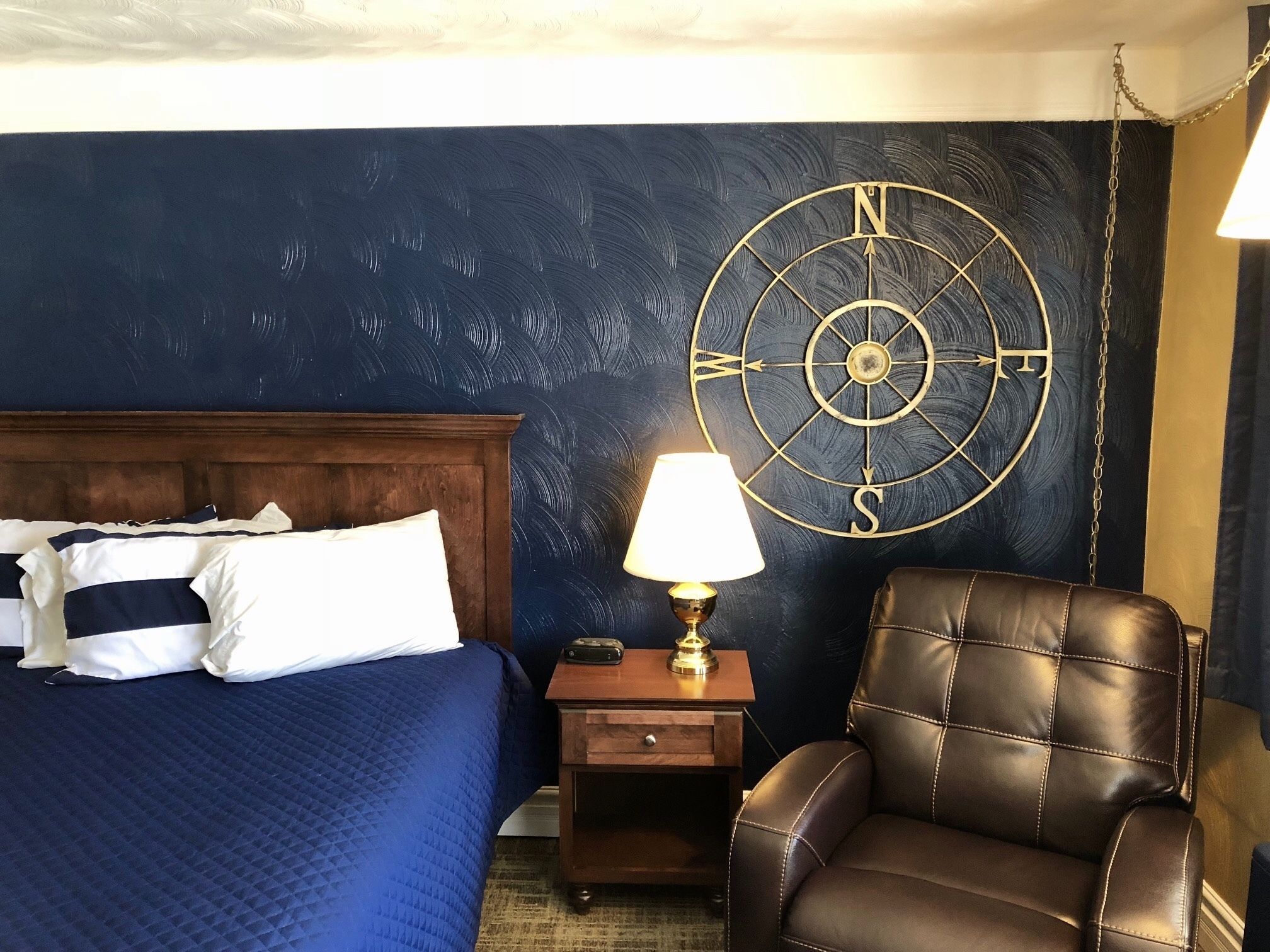 A hotel room with a bed, a leather armchair, a nightstand with a lamp, and a wall clock.