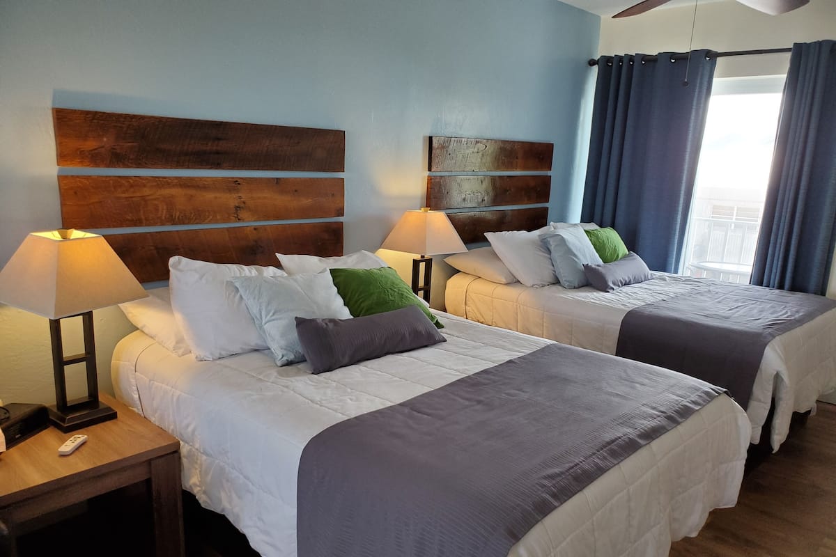 A hotel room with two beds, each with a wooden headboard, white bedding, and a bedside table with a lamp.