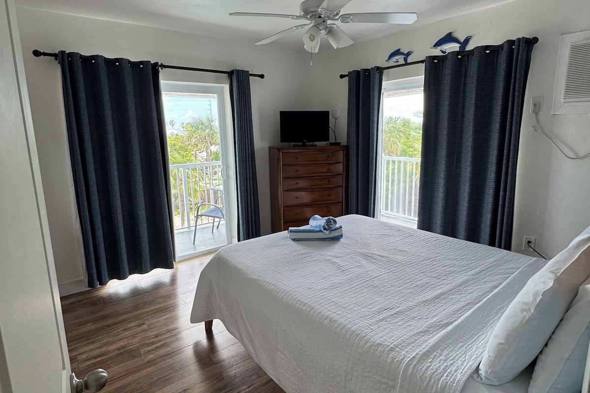 A bedroom with a bed, wooden dresser, ceiling fan, and a balcony with curtains.