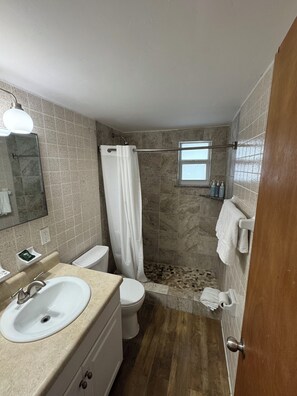 A bathroom with a sink, toilet, shower, and a small window.