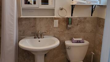 A bathroom with a pedestal sink, a toilet, and a mirror above the sink. There are towels on a shelf above the toilet.