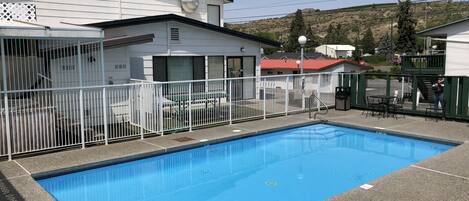 Una piscina con valla blanca, una zona de estar techada y un edificio con techo rojo al fondo.