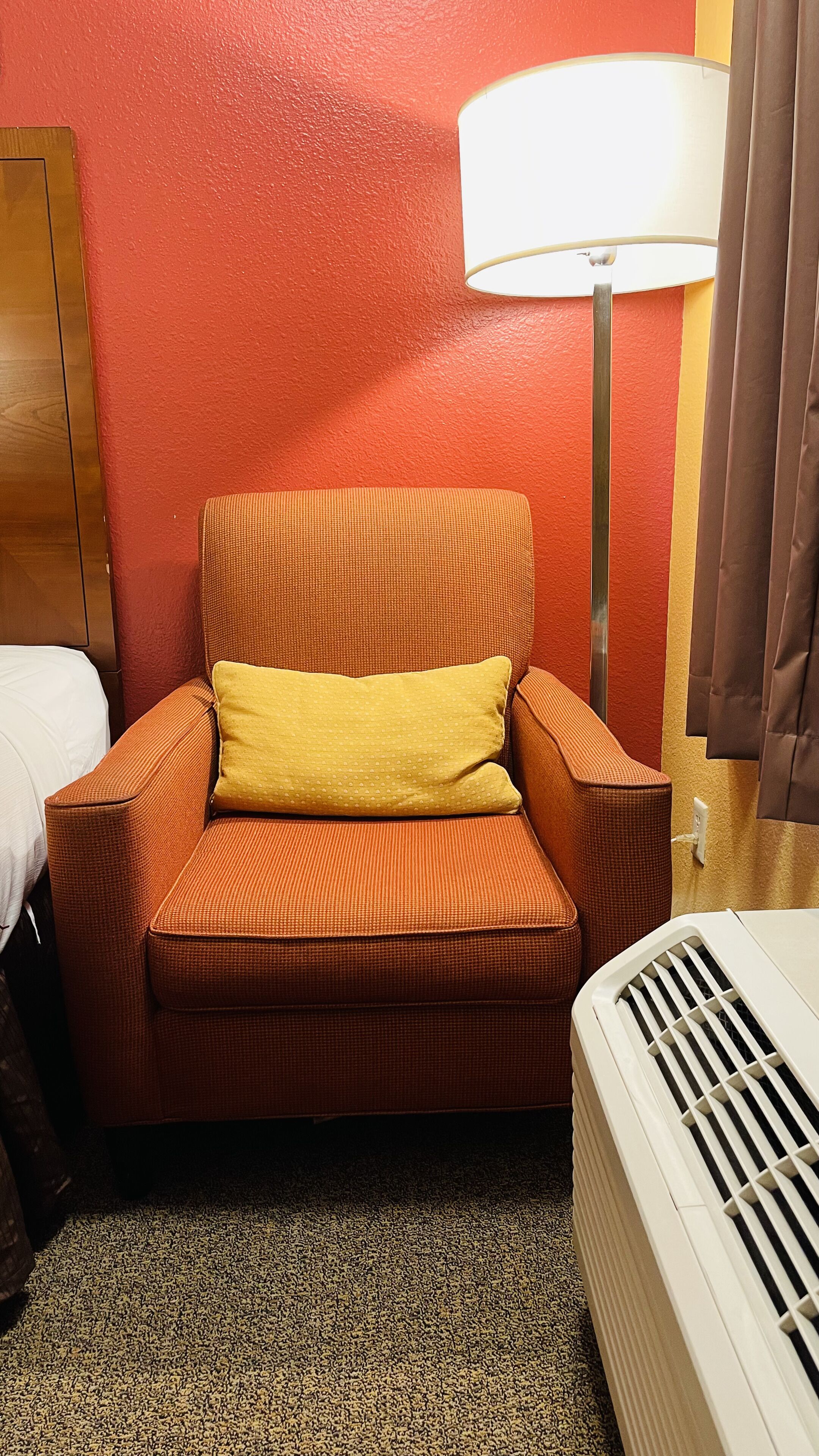 Une chambre d’hôtel avec un fauteuil orange, un coussin jaune, une table de chevet blanche, une lampe et un radiateur blanc.