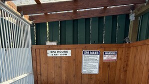 A hot tub with wooden walls and signs indicating spa hours, rules, and a warning about no lifeguard on duty.