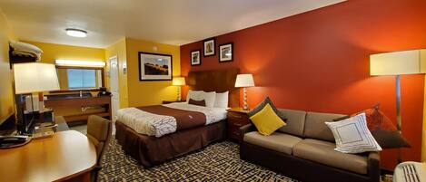 A hotel room with a bed, a sofa, a desk, and a chair. There are two lamps, a mirror, and framed pictures on the wall.