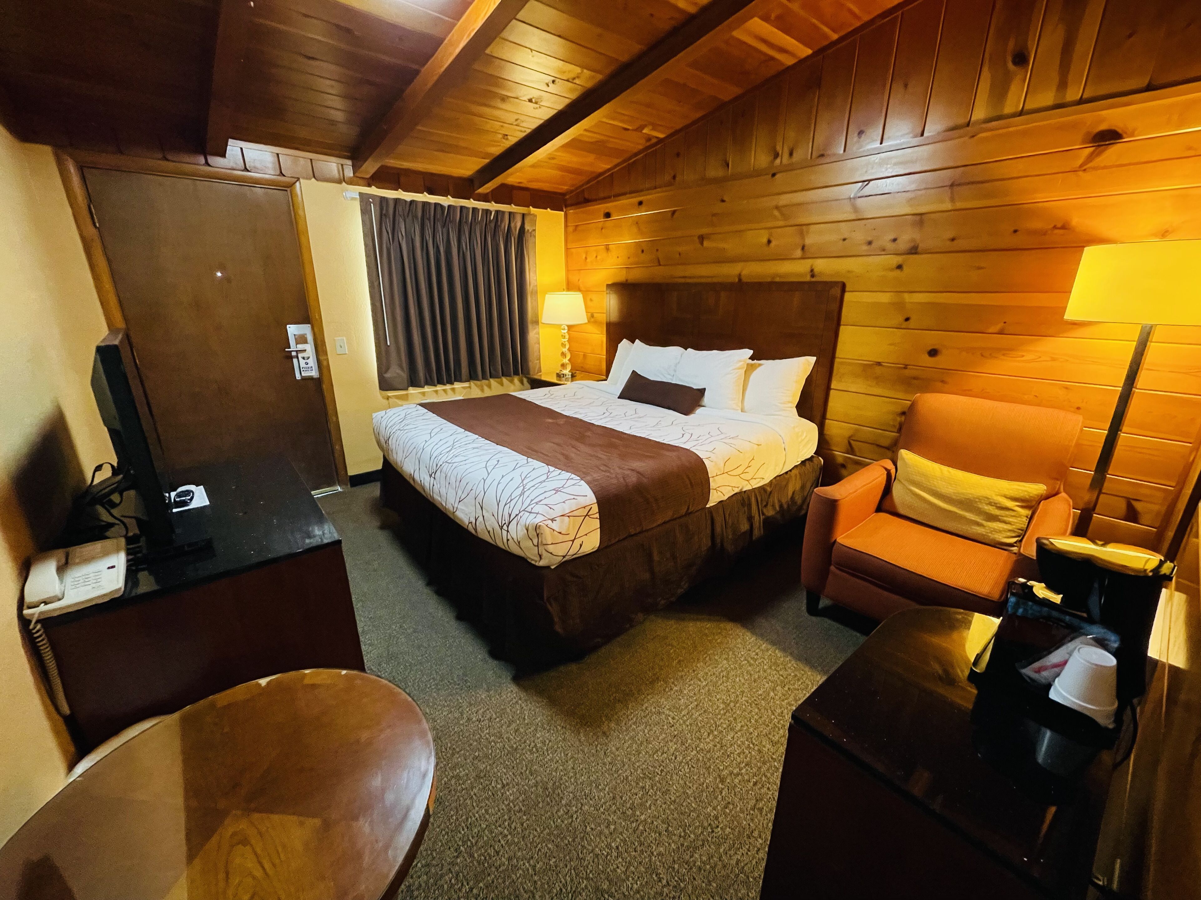 A wooden-paneled room with a bed, a chair, a desk, and a lamp.