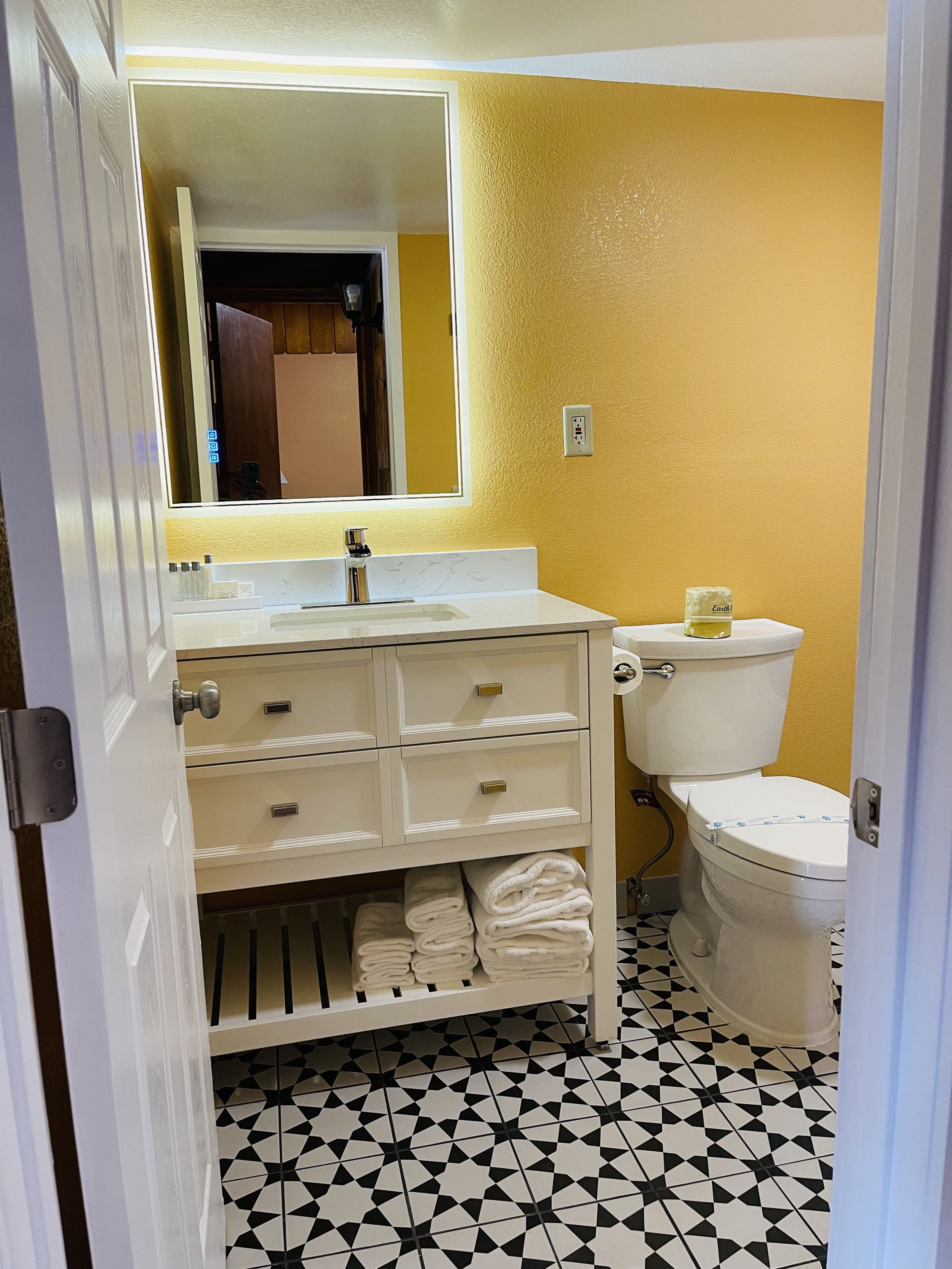 A bathroom with a white sink, mirror, and toilet. There are towels on a shelf below the sink. The floor has a black and white geometric pattern.
