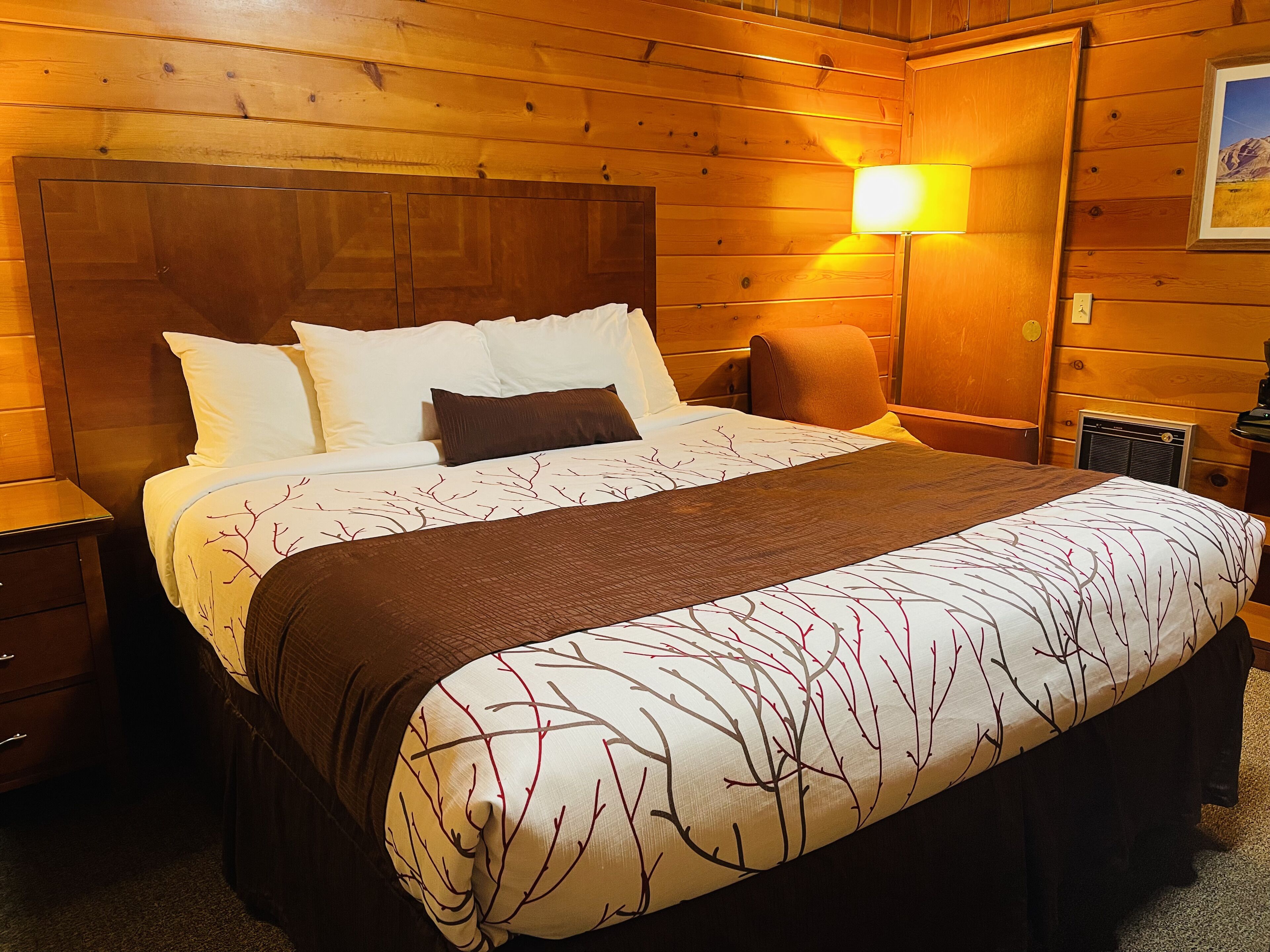 A wooden-paneled room with a bed, a nightstand, a lamp, and a framed picture on the wall.