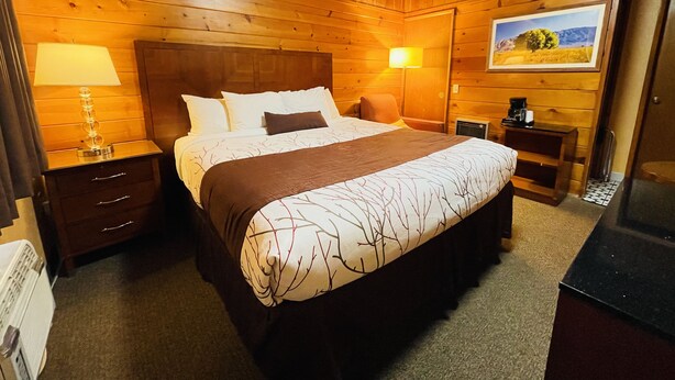 A wooden-paneled bedroom with a large bed, two bedside tables, a nightstand, a lamp, a framed picture on the wall, and a radiator.