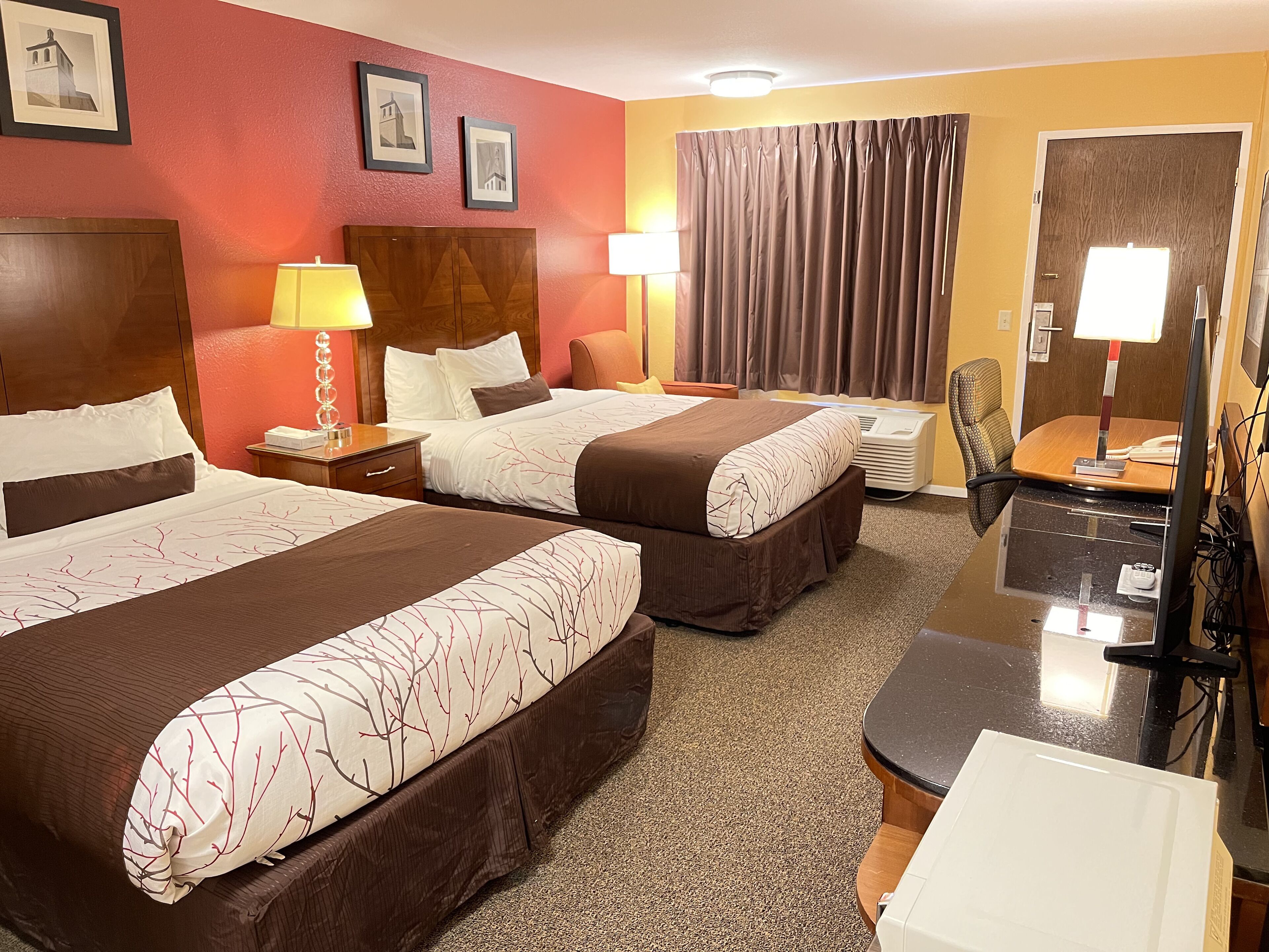 Une chambre d’hôtel avec deux lits, un bureau, une chaise, une lampe et une grande fenêtre avec des rideaux.