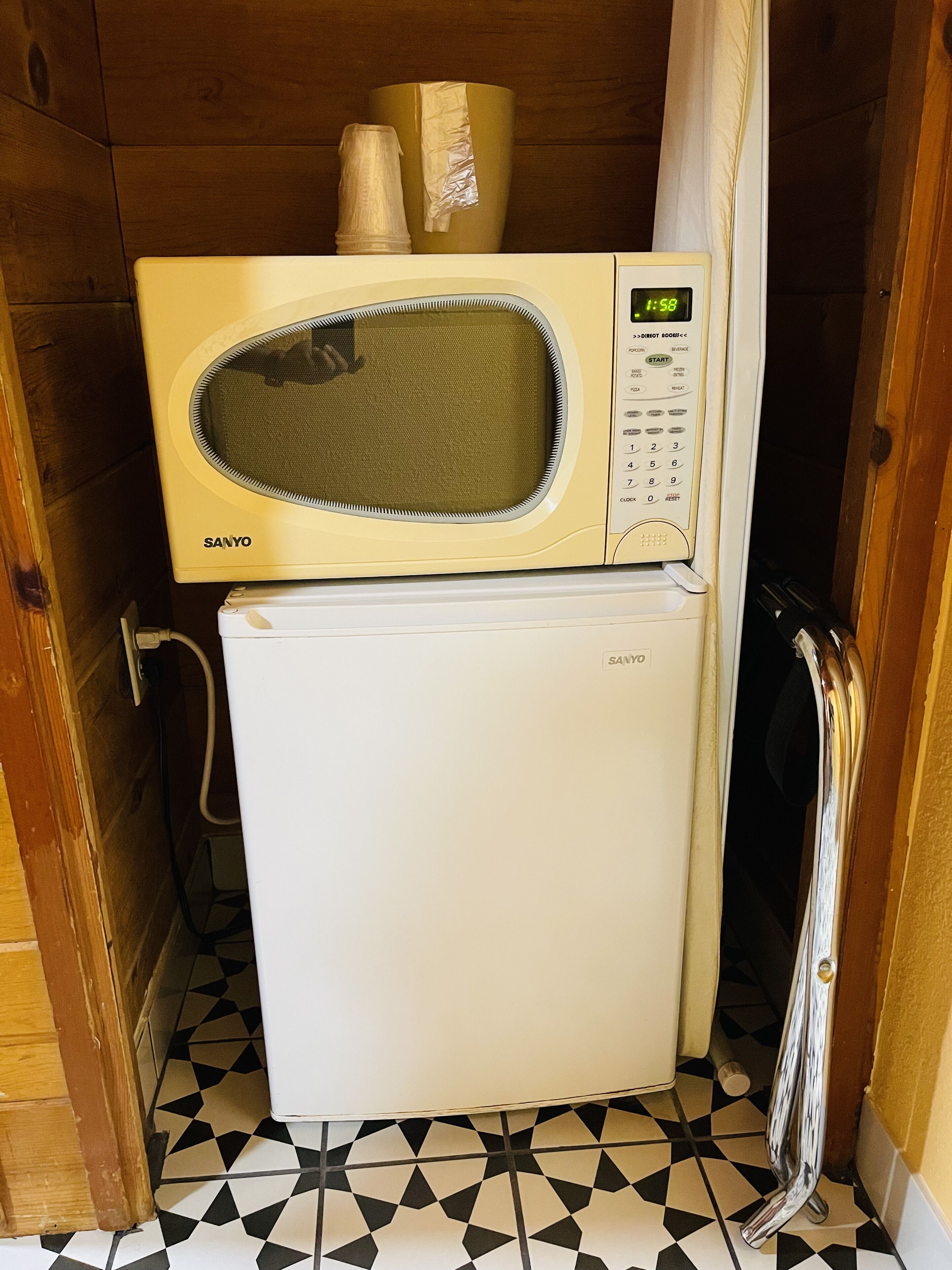 Een kleine keuken met een magnetron en een koelkast, houten wanden en een geruit vloertje.