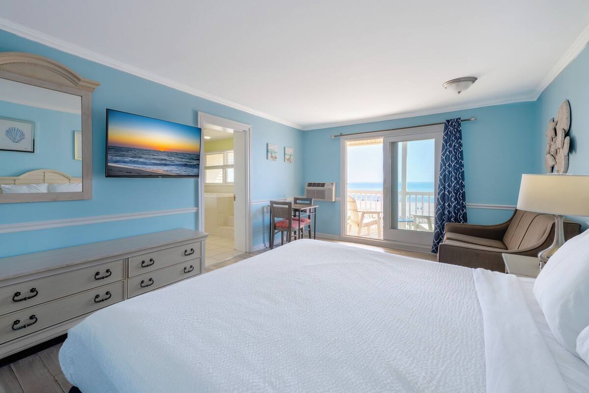 A hotel room with a large bed, a desk with a chair, a balcony with a view of the sea, and a painting of a sunset.