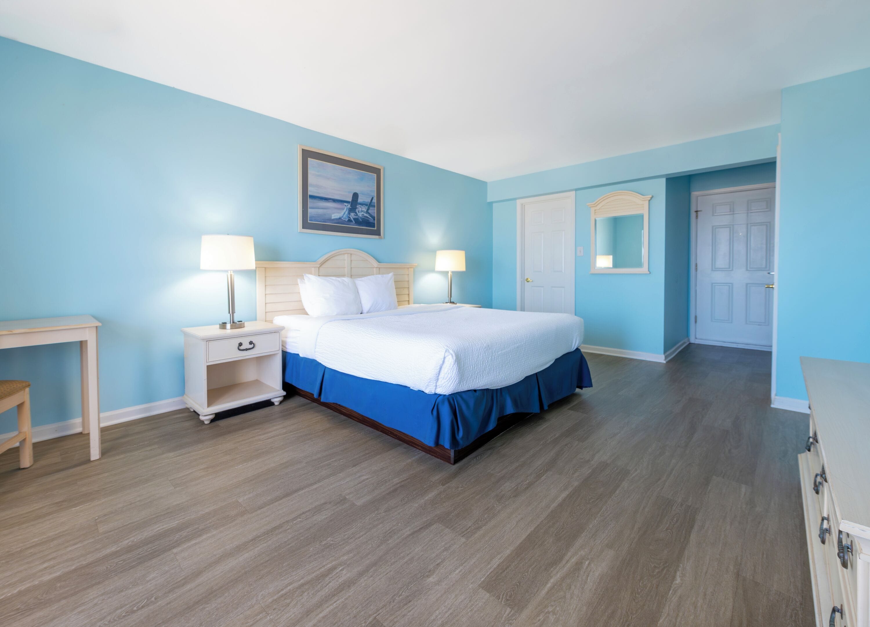 A bedroom with a bed, bedside tables, a nightstand, a dresser, a door, and a framed picture on the wall.