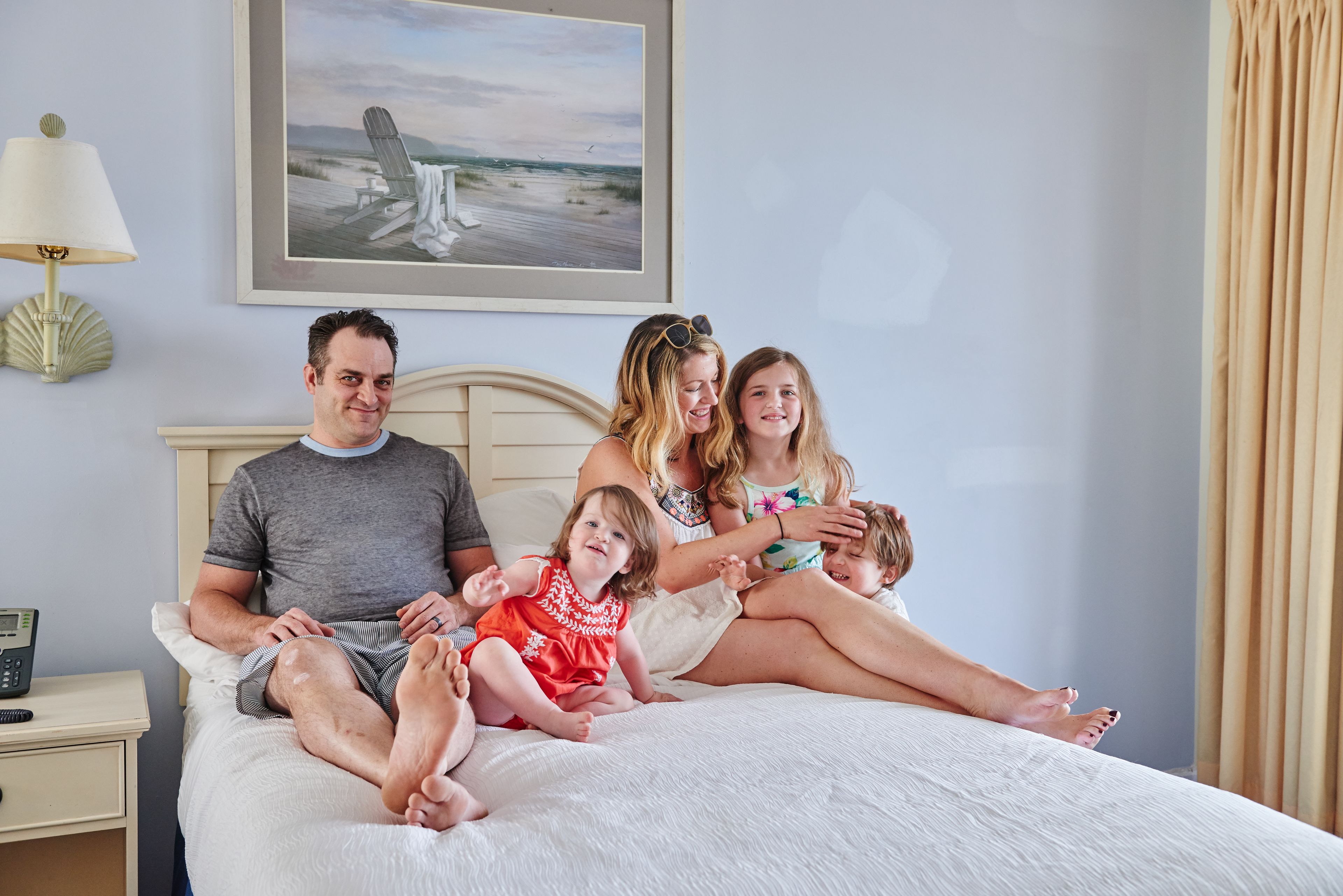 A family of four on a bed in a hotel room with a painting of a beach scene on the wall, a bedside lamp, and a nightstand with a phone.
