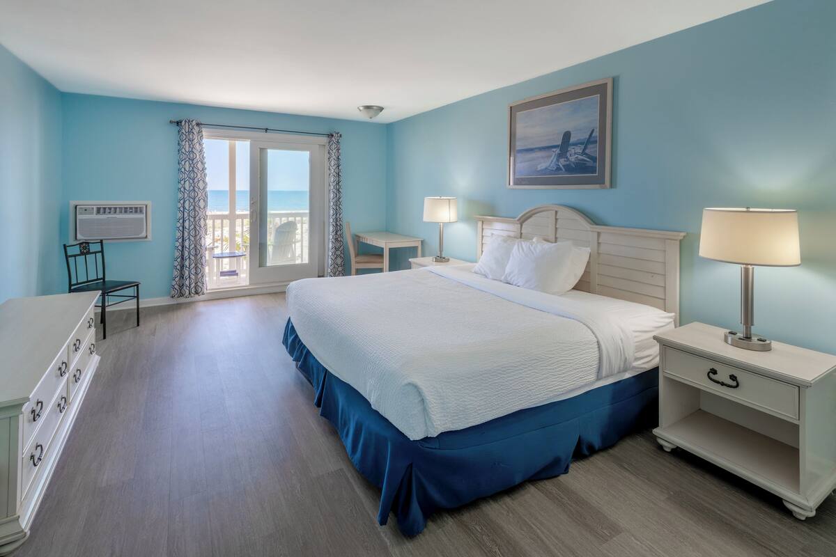 A hotel room with a bed, a desk, a chair, a nightstand, and a view of the ocean through a balcony door.