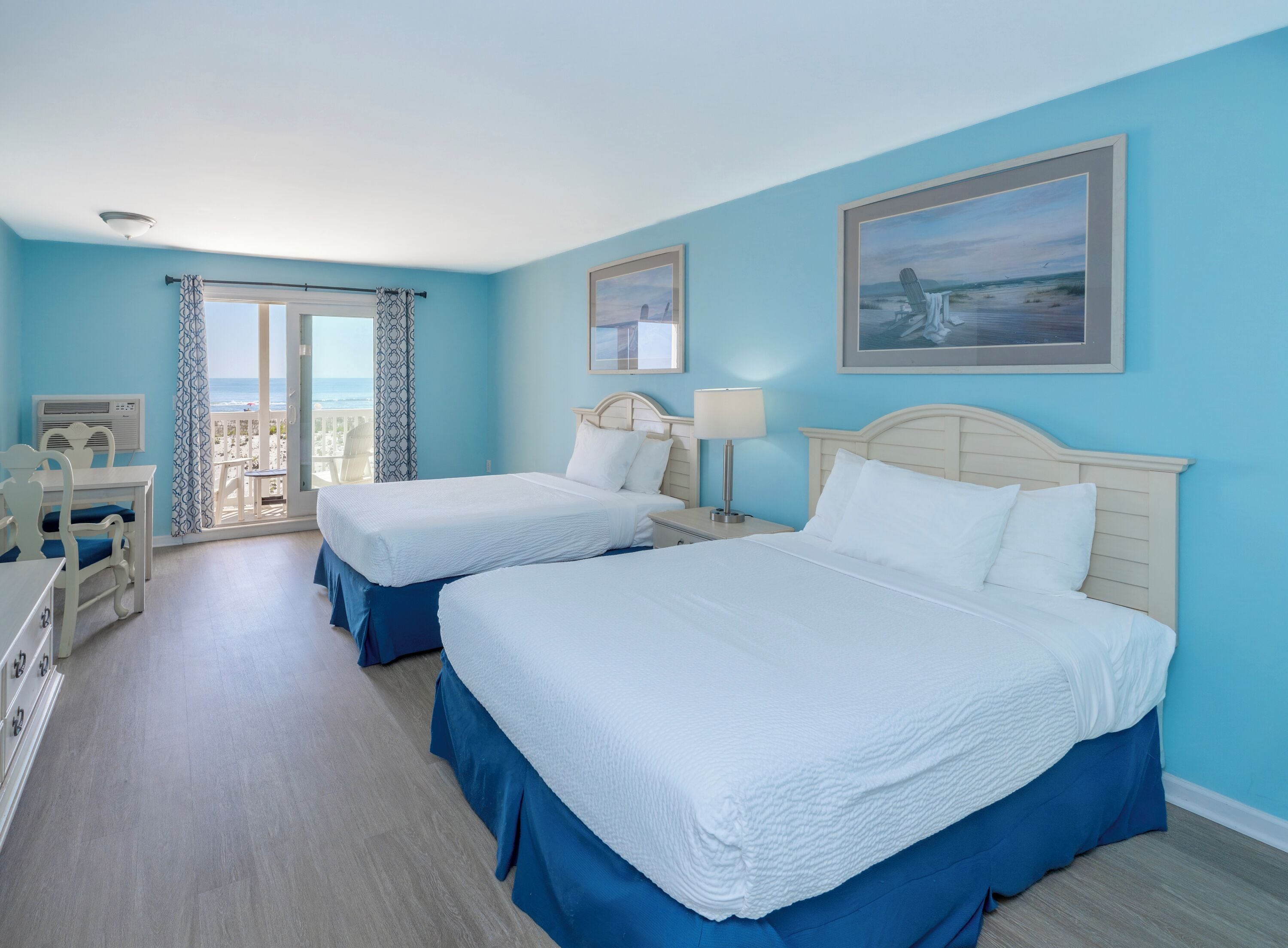 A hotel room with two beds, a balcony with a view of the sea, a table with chairs, and a ceiling fan.