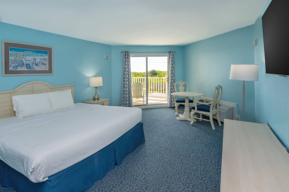 A hotel room with a large bed, a desk with two chairs, a balcony with a view, and a painting on the wall.