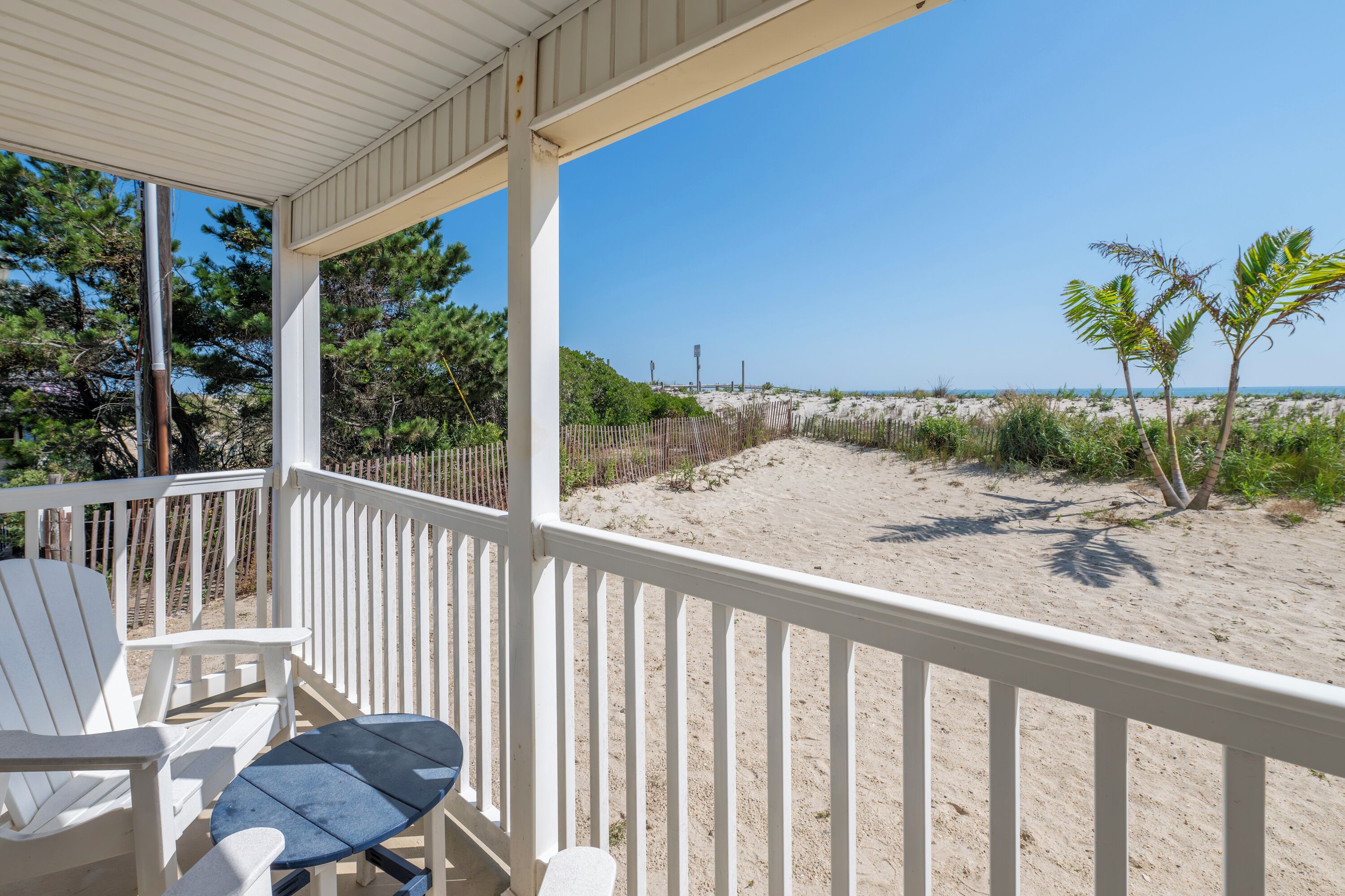 A covered outdoor area with white chairs and a table, overlooking a sandy beach with sparse vegetation and a clear sky.