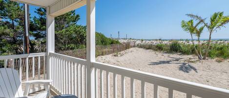 A covered outdoor area with white chairs and a table, overlooking a sandy beach with sparse vegetation and a clear sky.