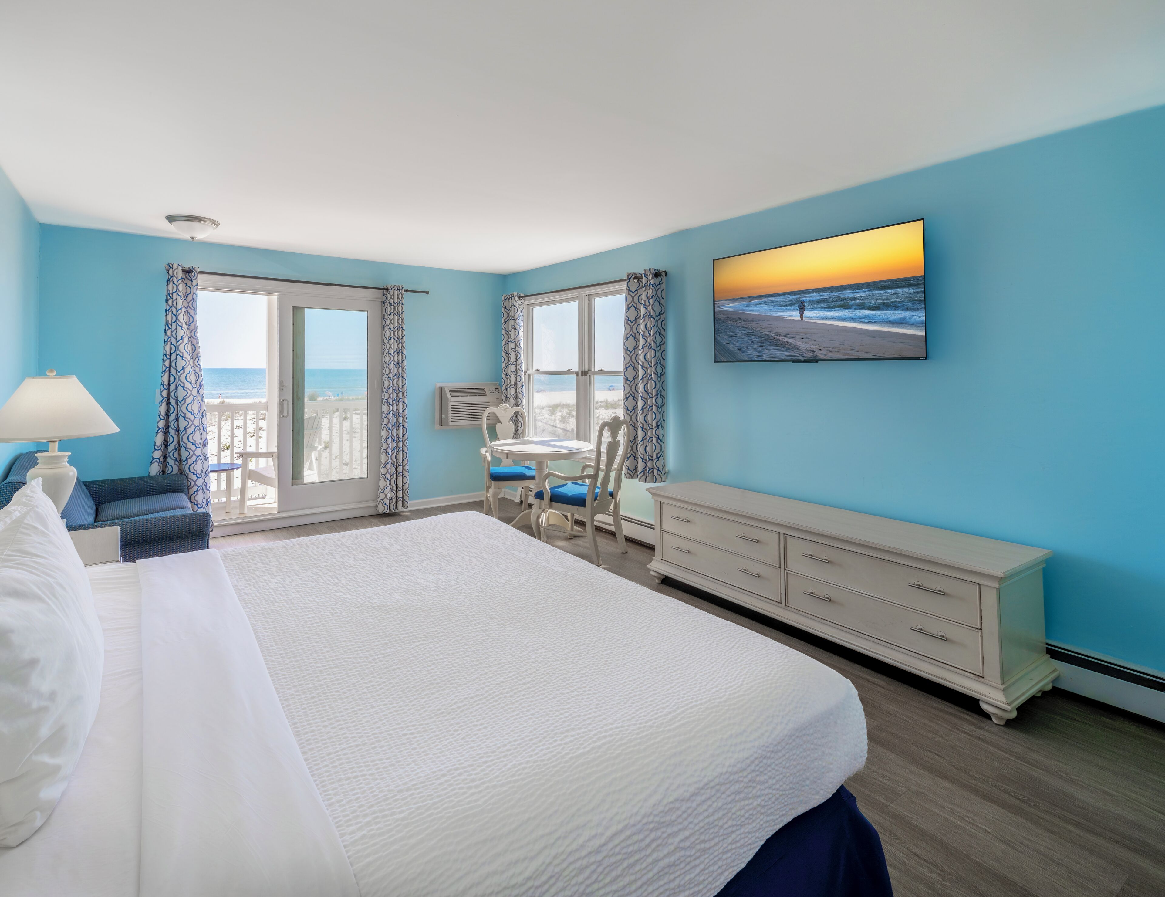 A hotel room with a large bed, a desk with two chairs, a dresser, and a view of the beach through the window.