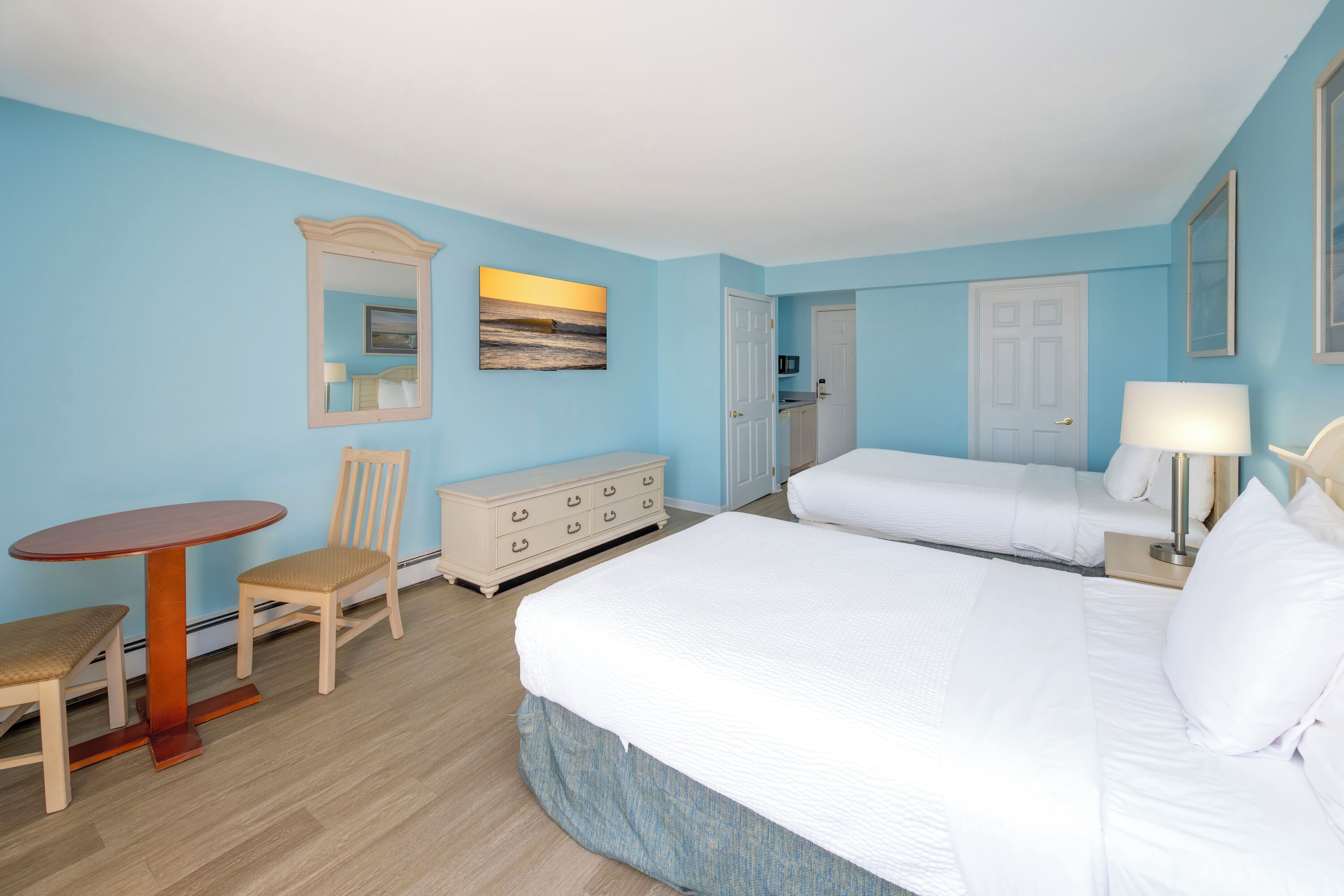 A hotel room with a large bed, a small table, a chair, a dresser, and a painting on the wall.