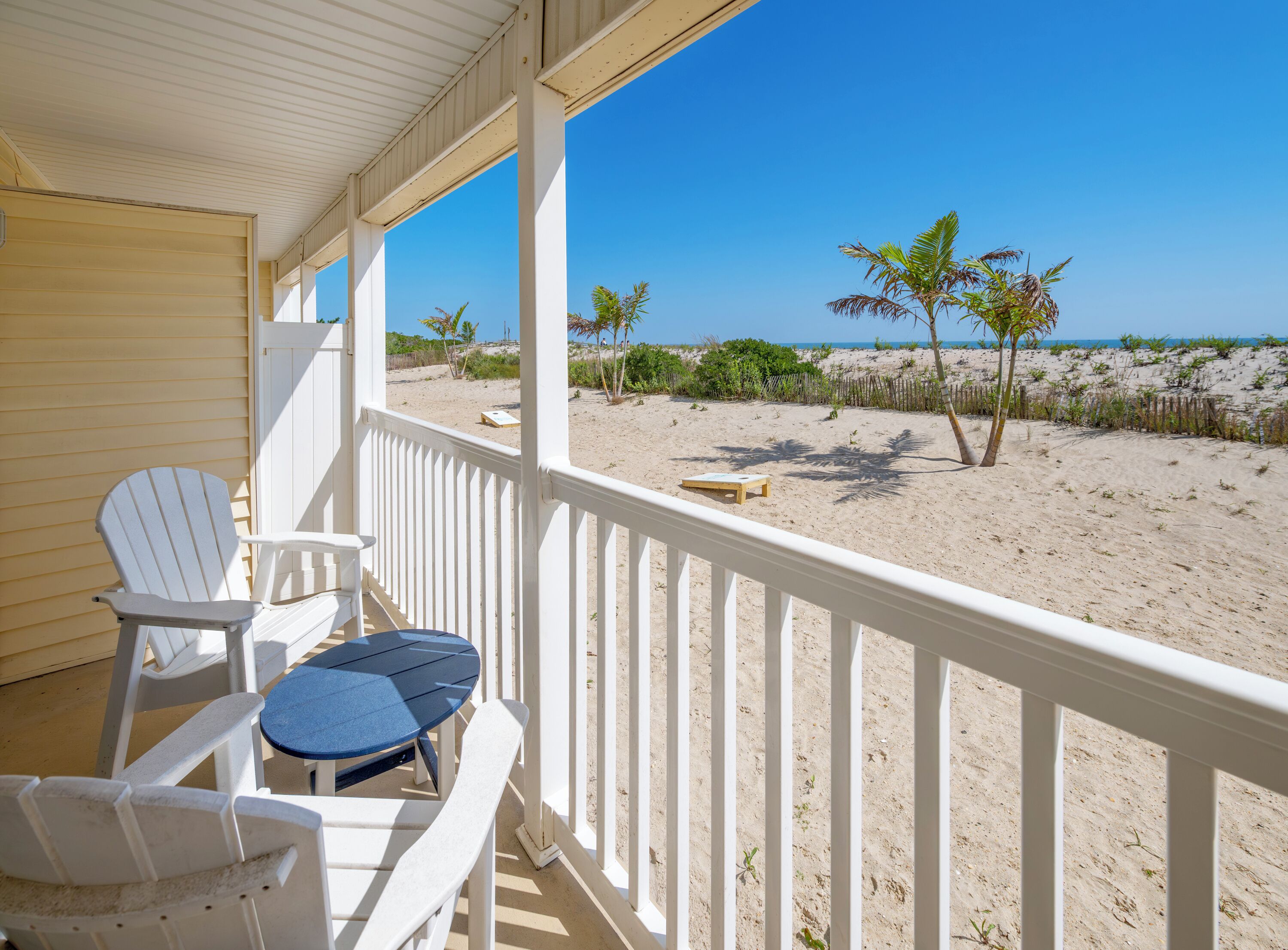 A covered porch with white Adirondack chairs and a small table, overlooking a sandy beach with sparse vegetation and a clear blue sky.