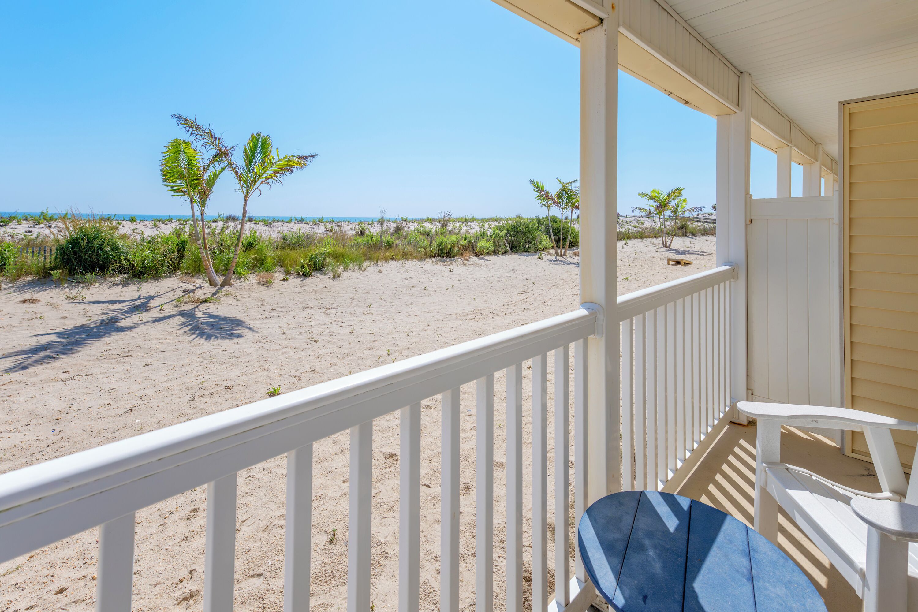 A beach view from a porch with a white railing, a blue table, and a wooden door.