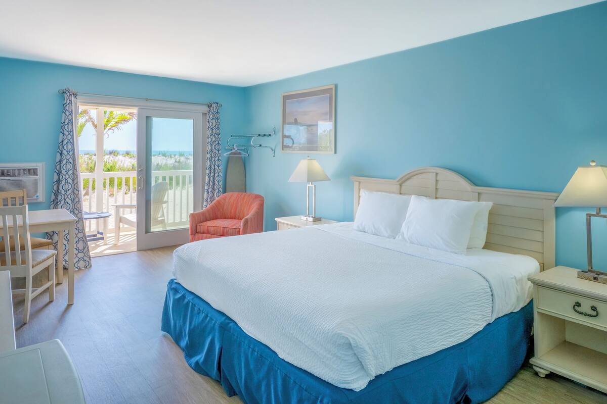 A bedroom with a large bed, a chair, a nightstand, a lamp, and a view of the outdoors.