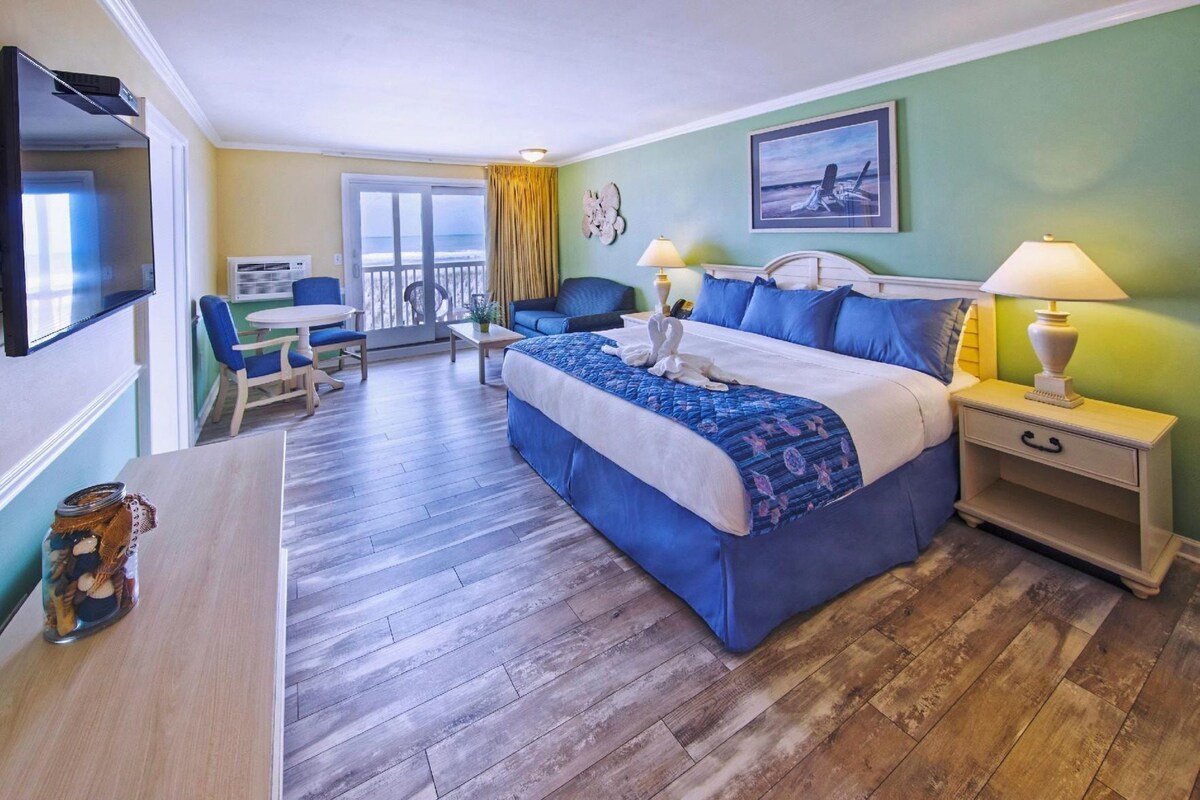 Habitación de hotel con cama, escritorio, silla, televisor y balcón con vistas.