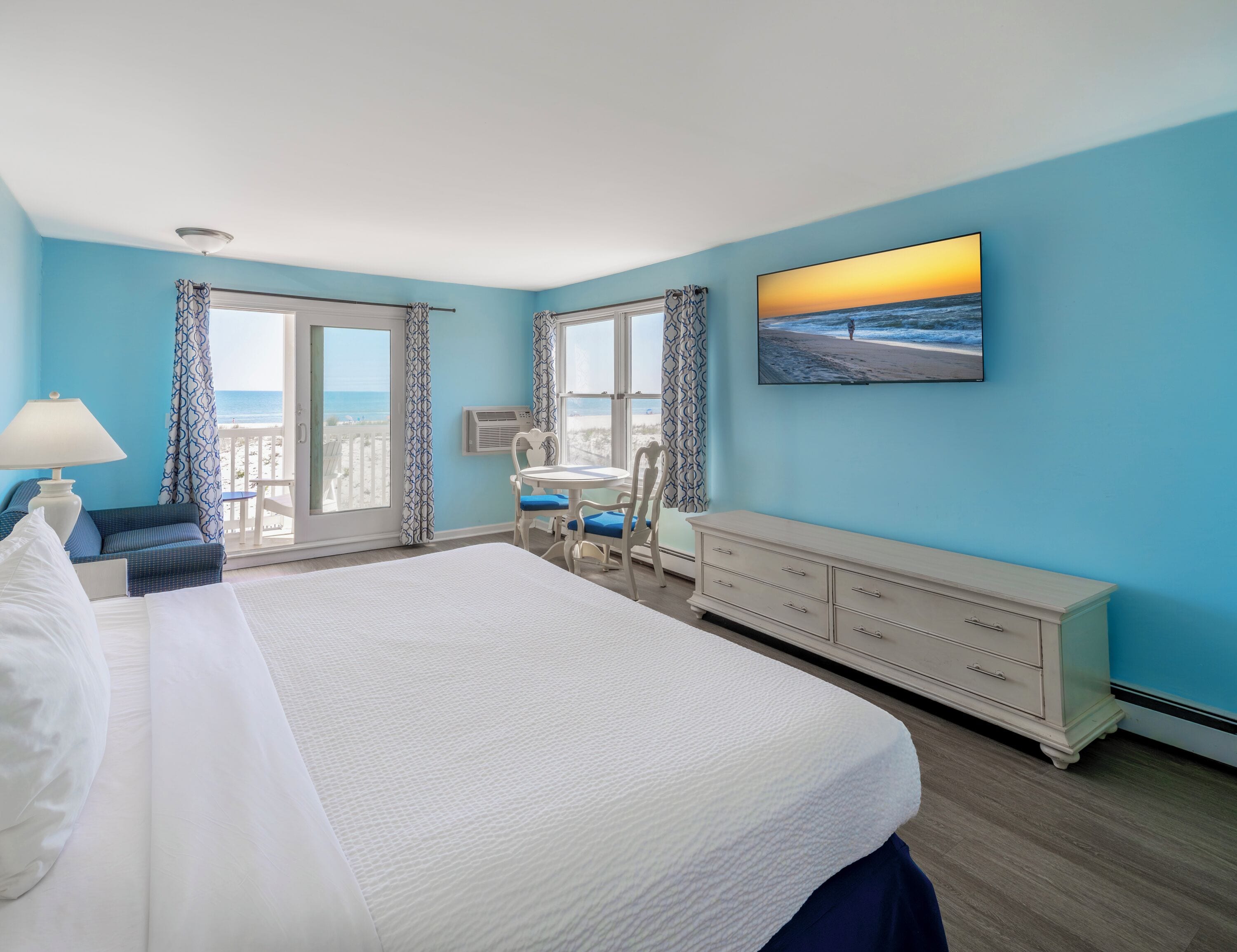 A hotel room with a large bed, a desk with two chairs, a dresser, and a large window with a view of the beach.