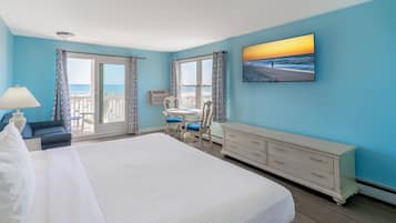 A hotel room with a large bed, a desk with two chairs, a dresser, and a large window with a view of the beach.
