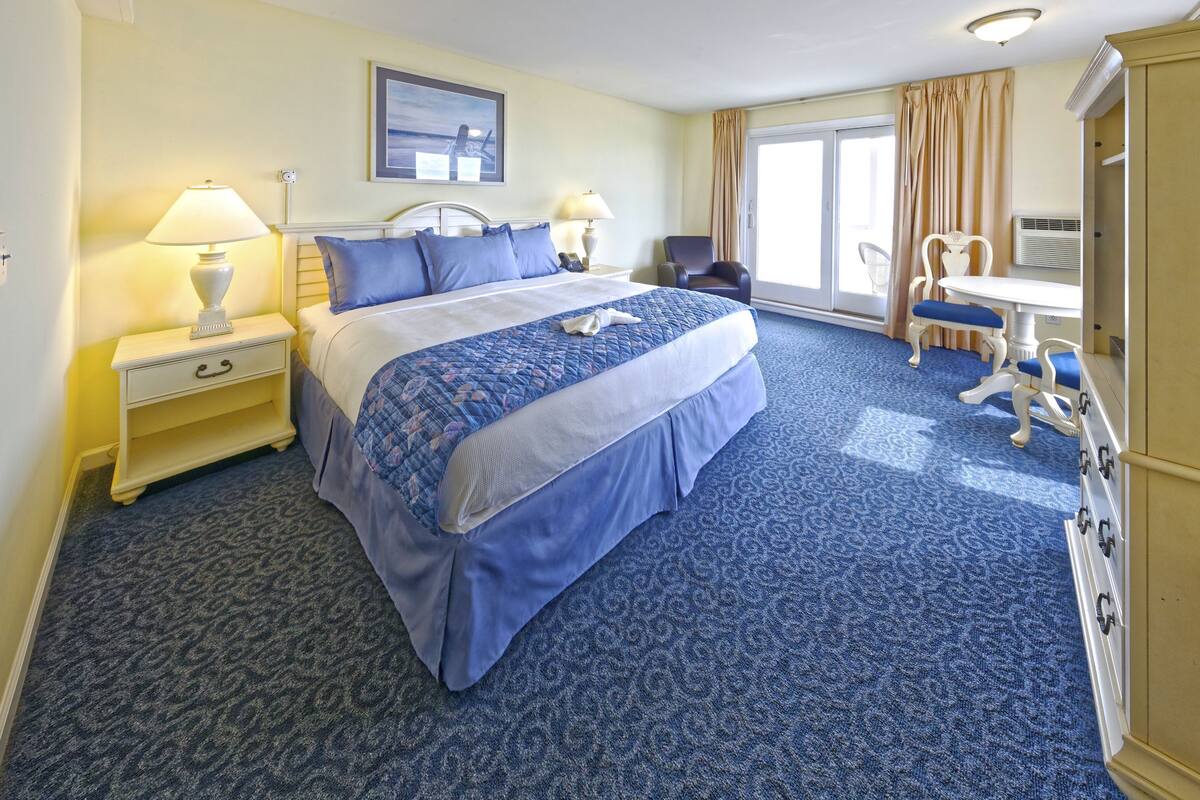 A hotel room with a large bed, two bedside tables with lamps, a desk with a chair, and a large window with curtains.