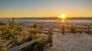 Escena de playa con una valla de madera, arena y un atardecer sobre el océano.