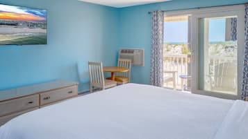 Un dormitorio con cama, escritorio y silla. Hay una ventana con vistas a la playa y un balcón con sillas.