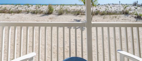 A beach view from a covered deck with white railings, two white Adirondack chairs, and a round blue table.