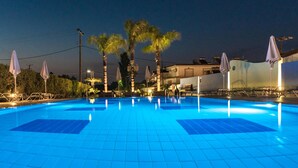 Una piscina con suelo de baldosas, rodeada de palmeras y sombrillas, con edificios al fondo.