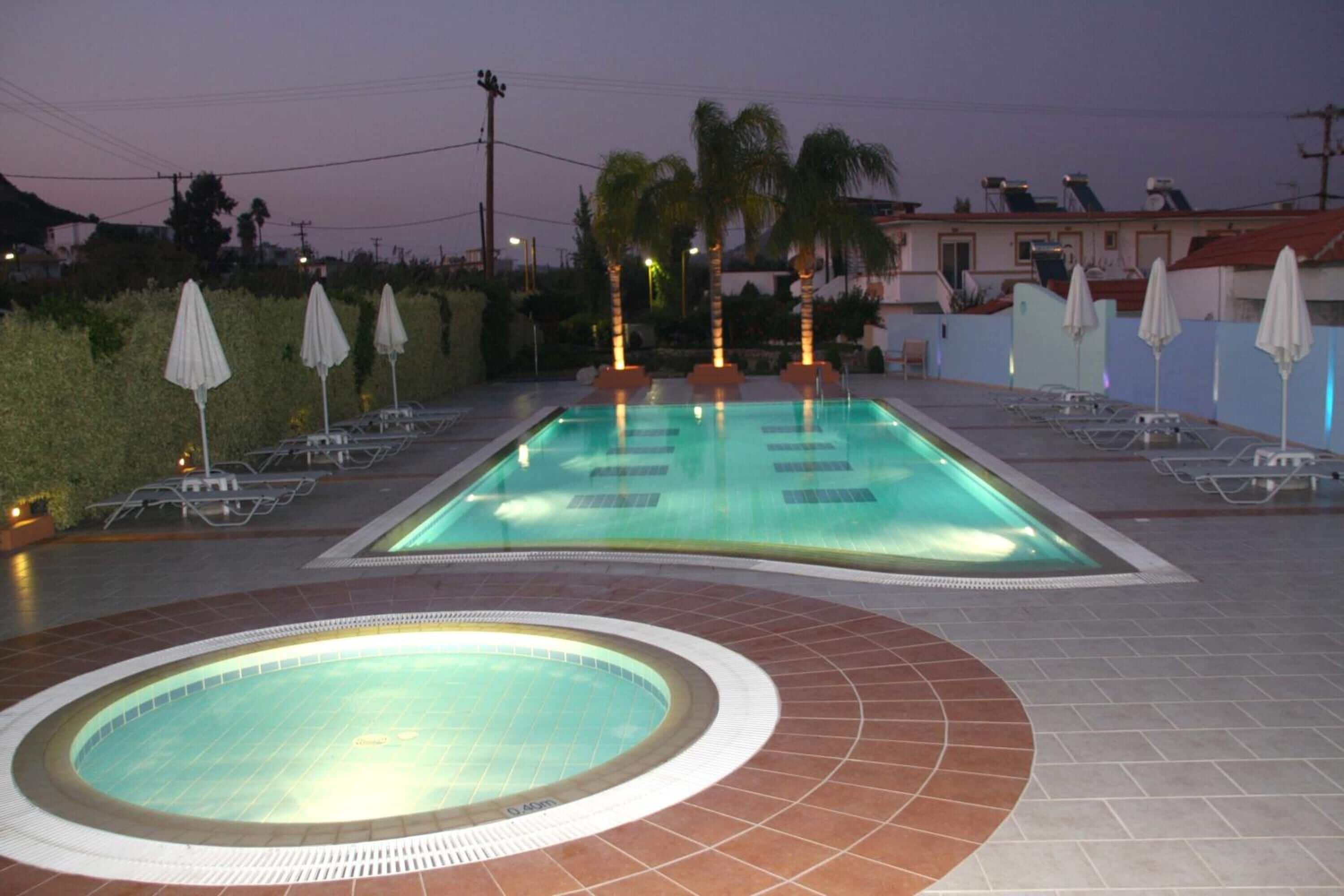 Une piscine entourée de palmiers, avec des chaises longues et des parasols, et un bâtiment visible au loin.