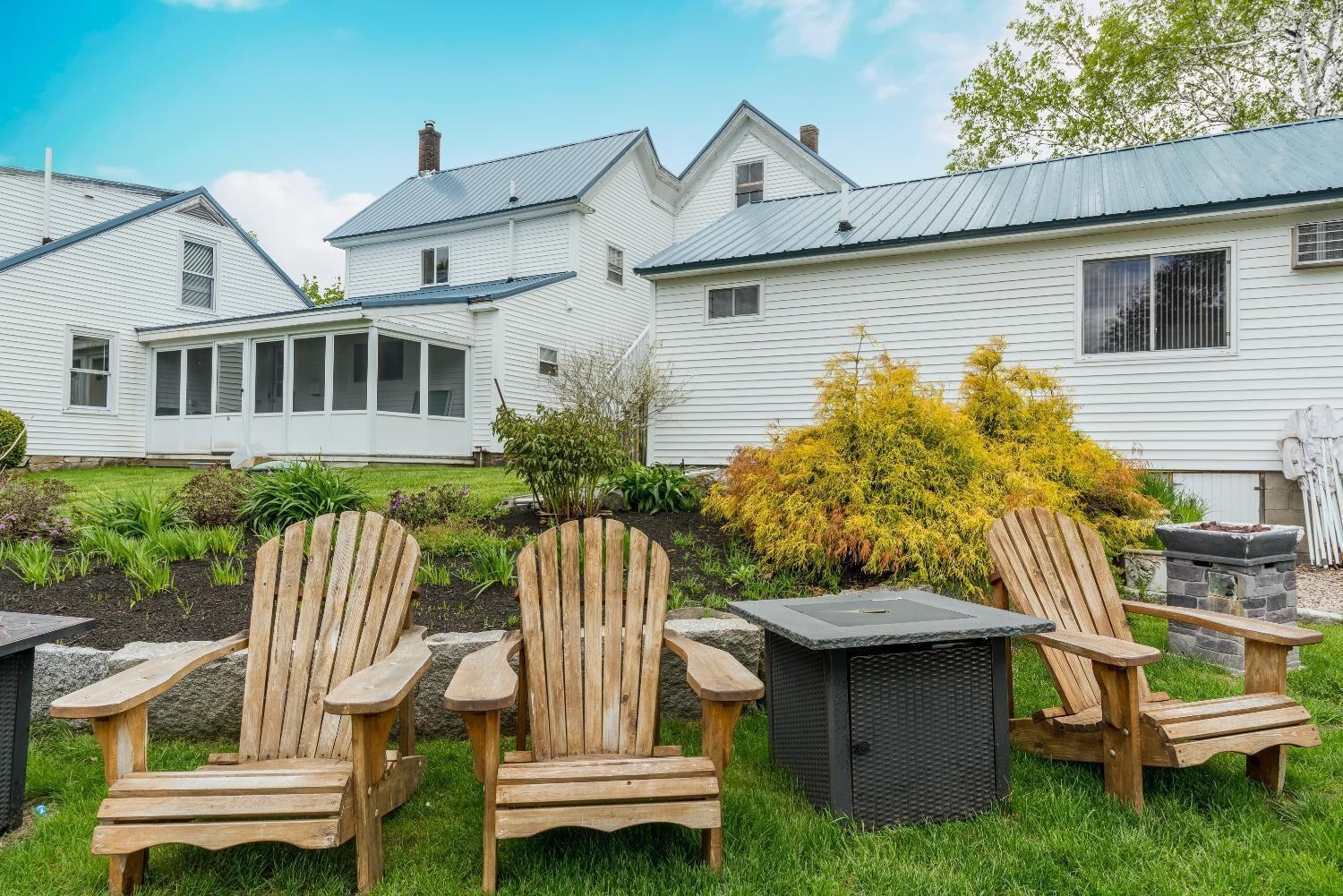 A row of Adirondack chairs in a garden area with a stone fire pit and a small table.