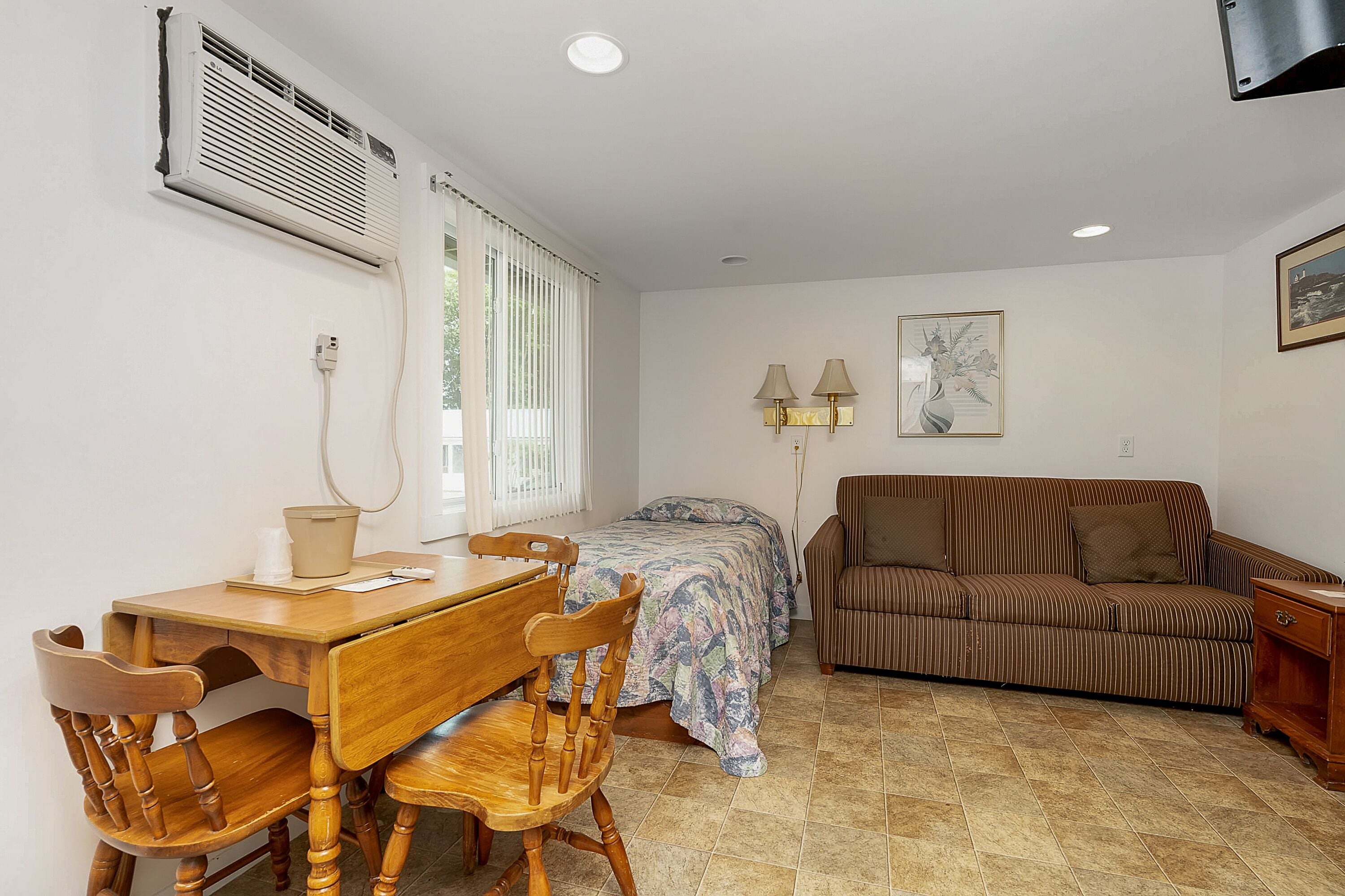 A compact room with a bed, a sofa, a dining table with chairs, and an air conditioning unit.
