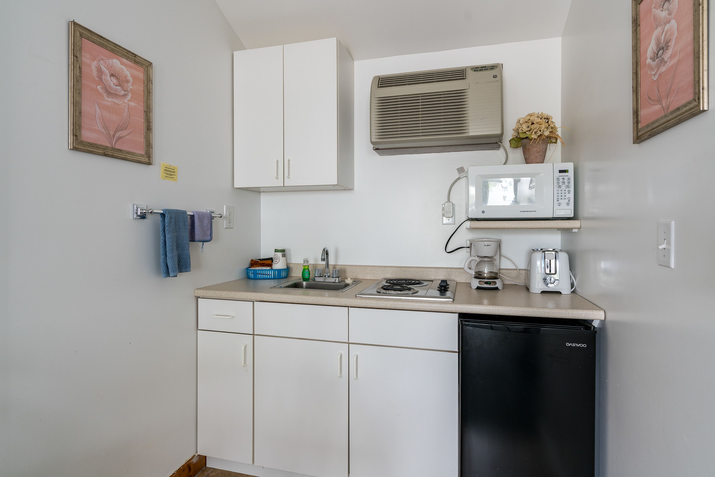Una cocina compacta con armarios blancos, un lavavajillas negro, microondas, cafetera y un lavabo pequeño.