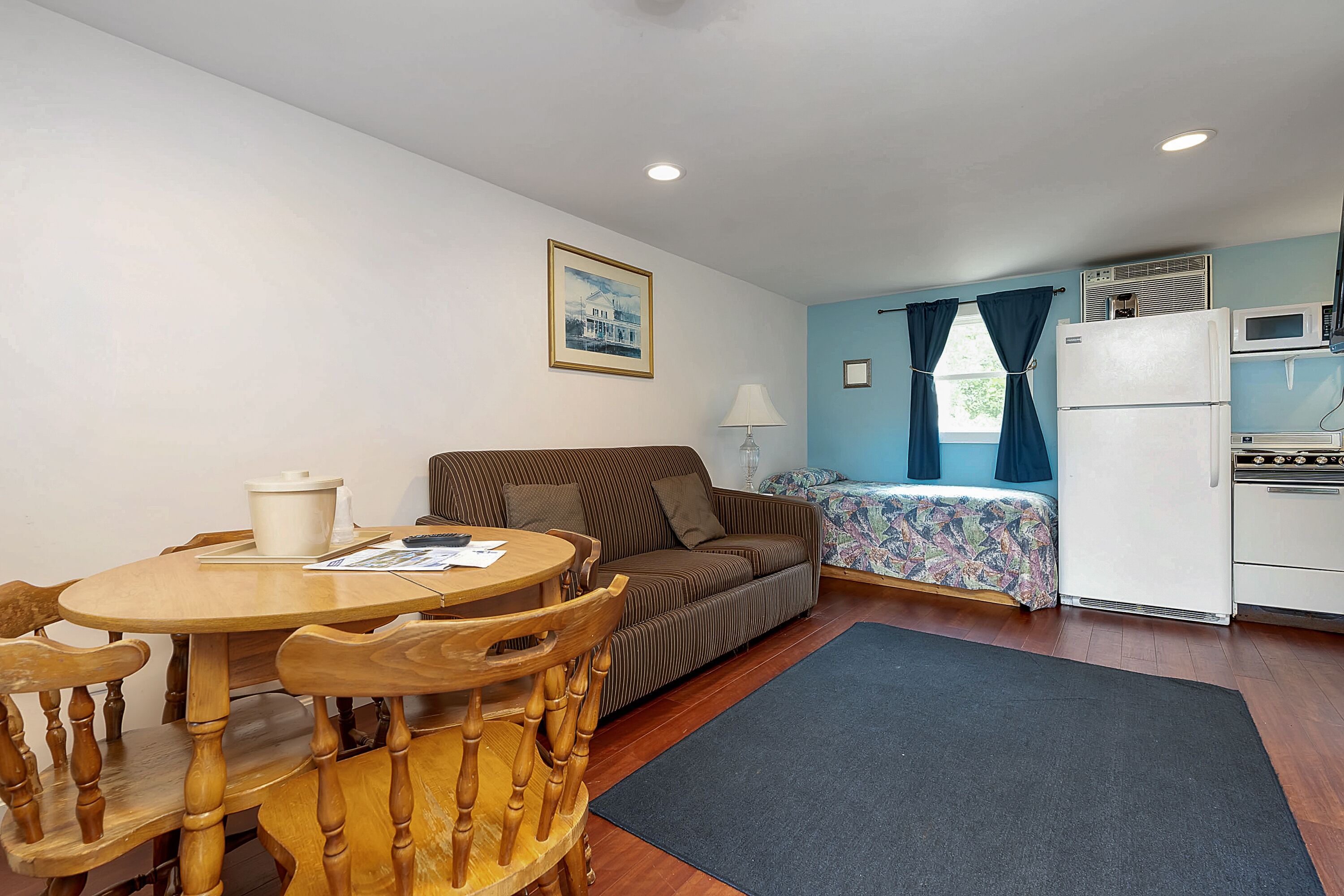 Un espacio habitable compacto con una mesa de comedor, sillas de madera, un sofá, una cama con colcha colorida, una kitchenette con microondas y estufa, y una ventana con cortinas.