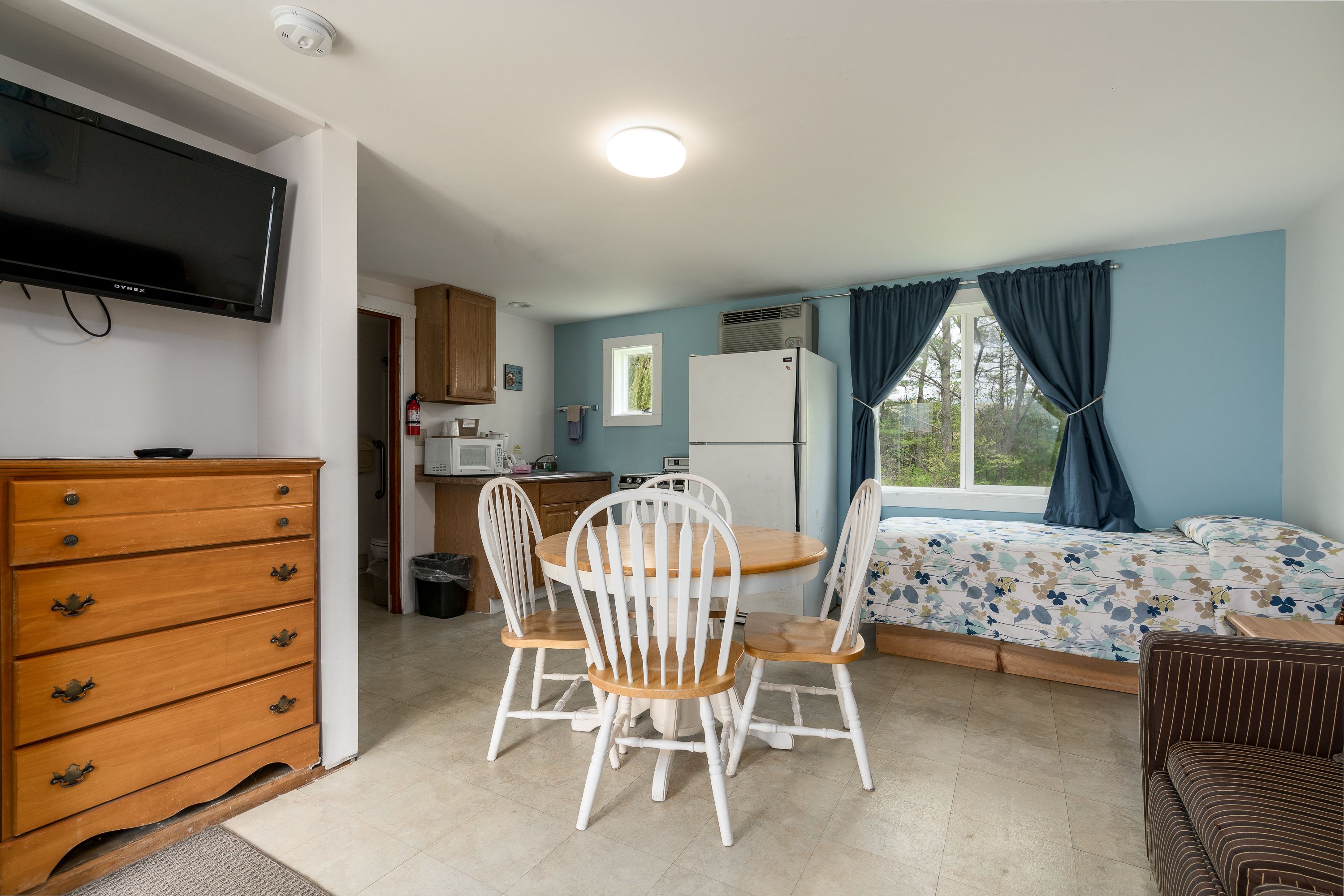 A compact room with a wooden dresser, a television, a dining table with white chairs, a bed with a floral bedspread, and a window with curtains.