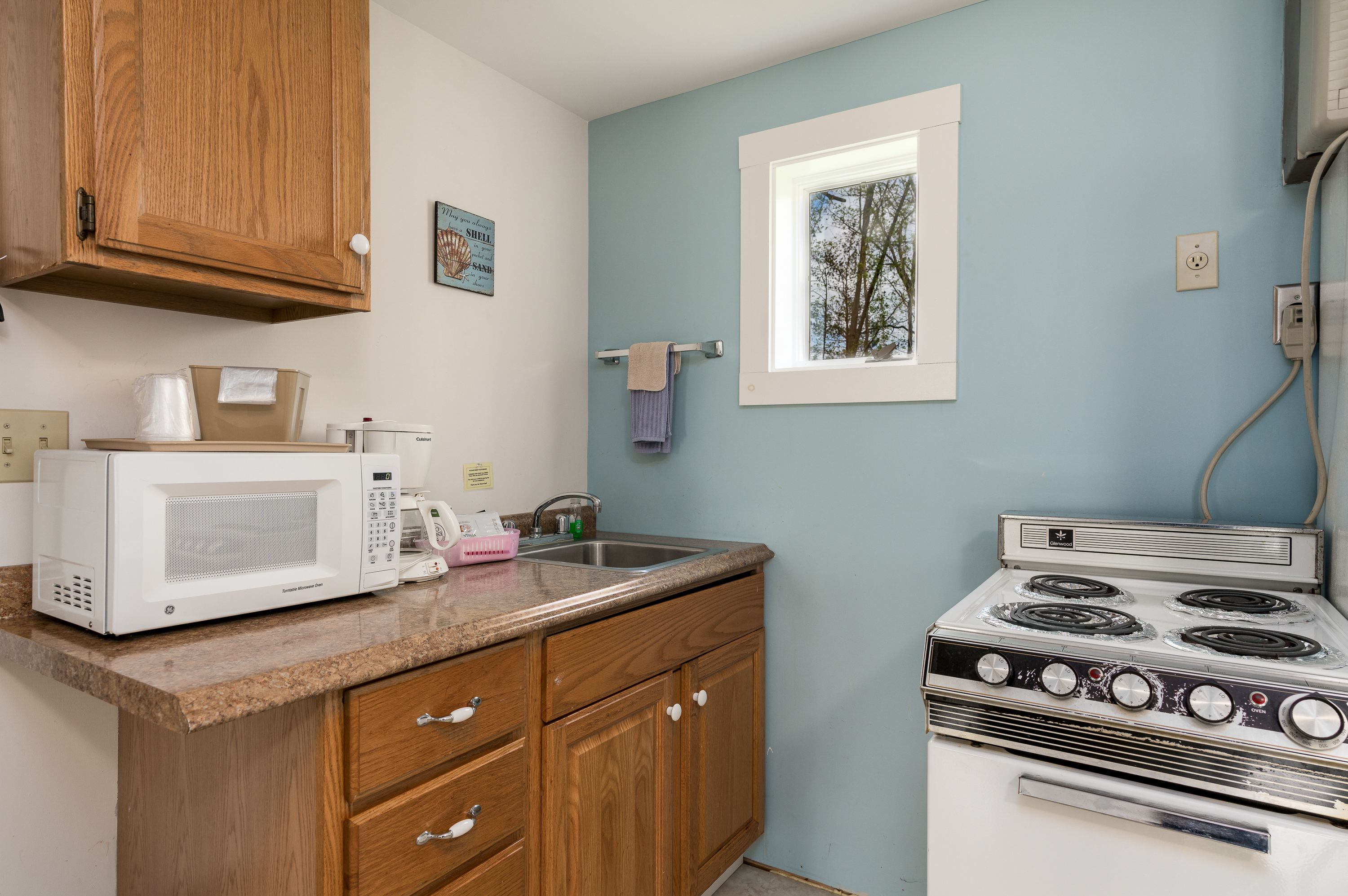 A kitchen with wooden cabinets, a white stove, a microwave, and a window with a view of trees.