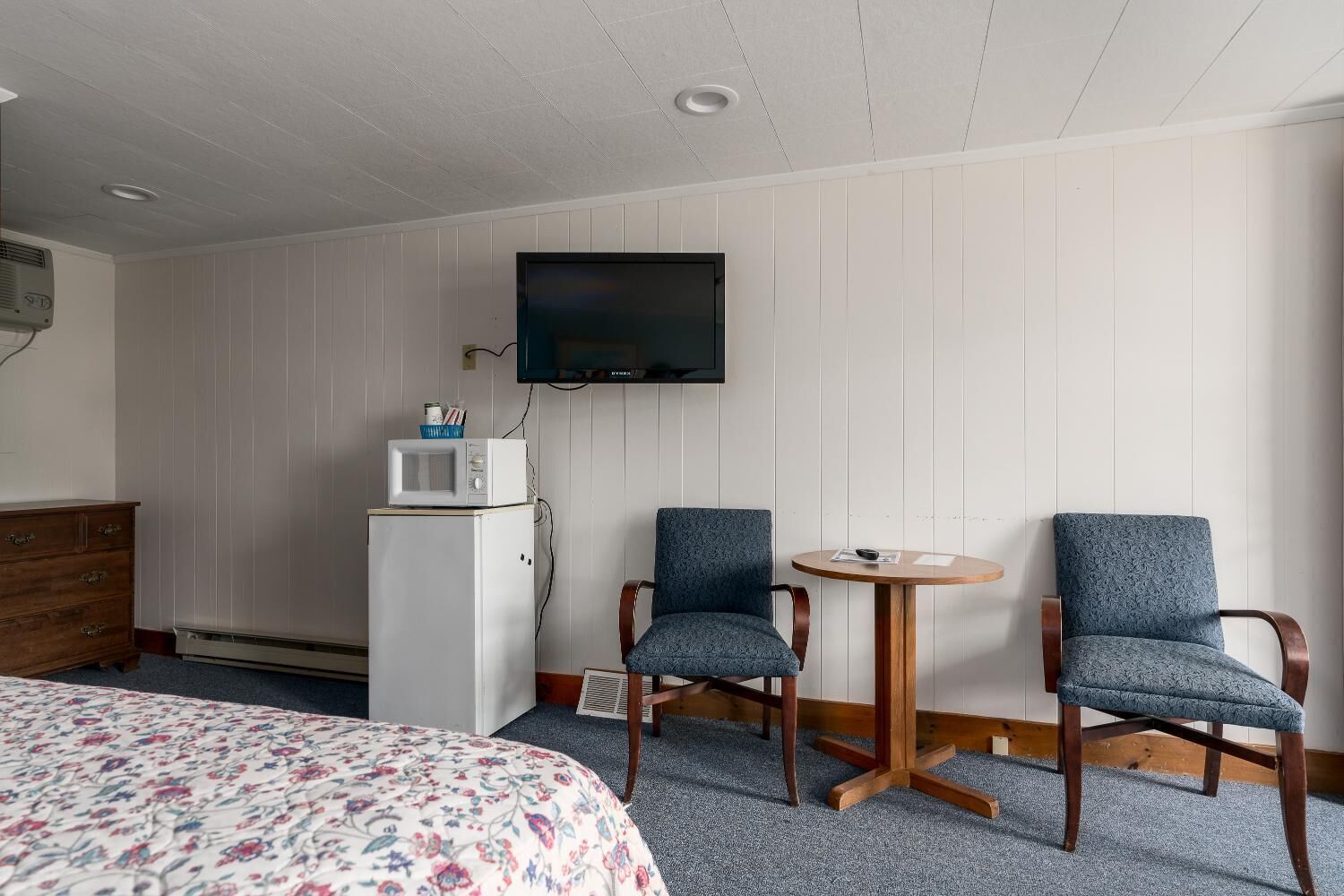 A hotel room with a bed, two armchairs, a small table, a microwave, and a television mounted on the wall.