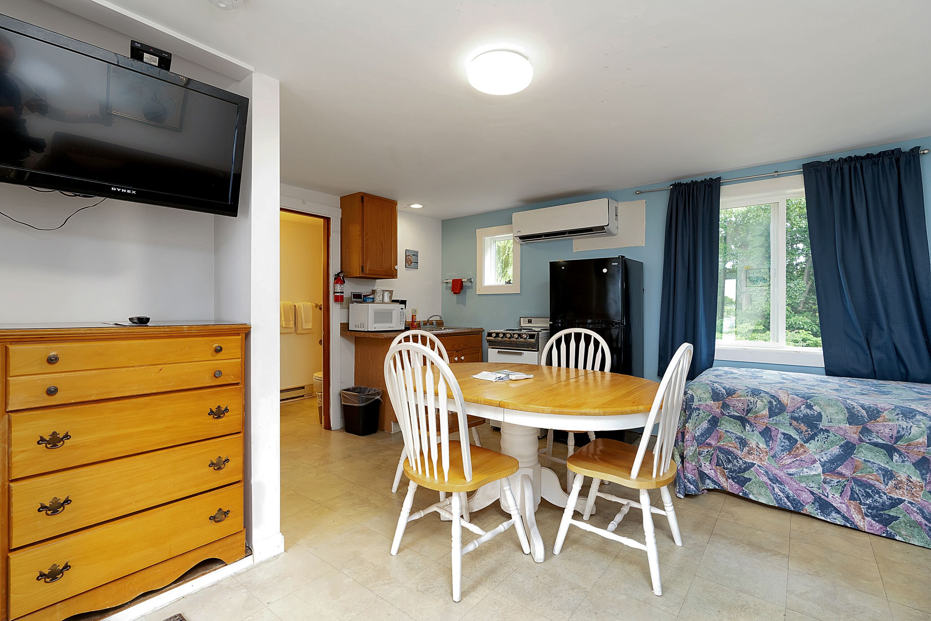 A compact kitchen with a microwave, stove, and refrigerator. There is a dining table with four chairs and a TV mounted on the wall.
