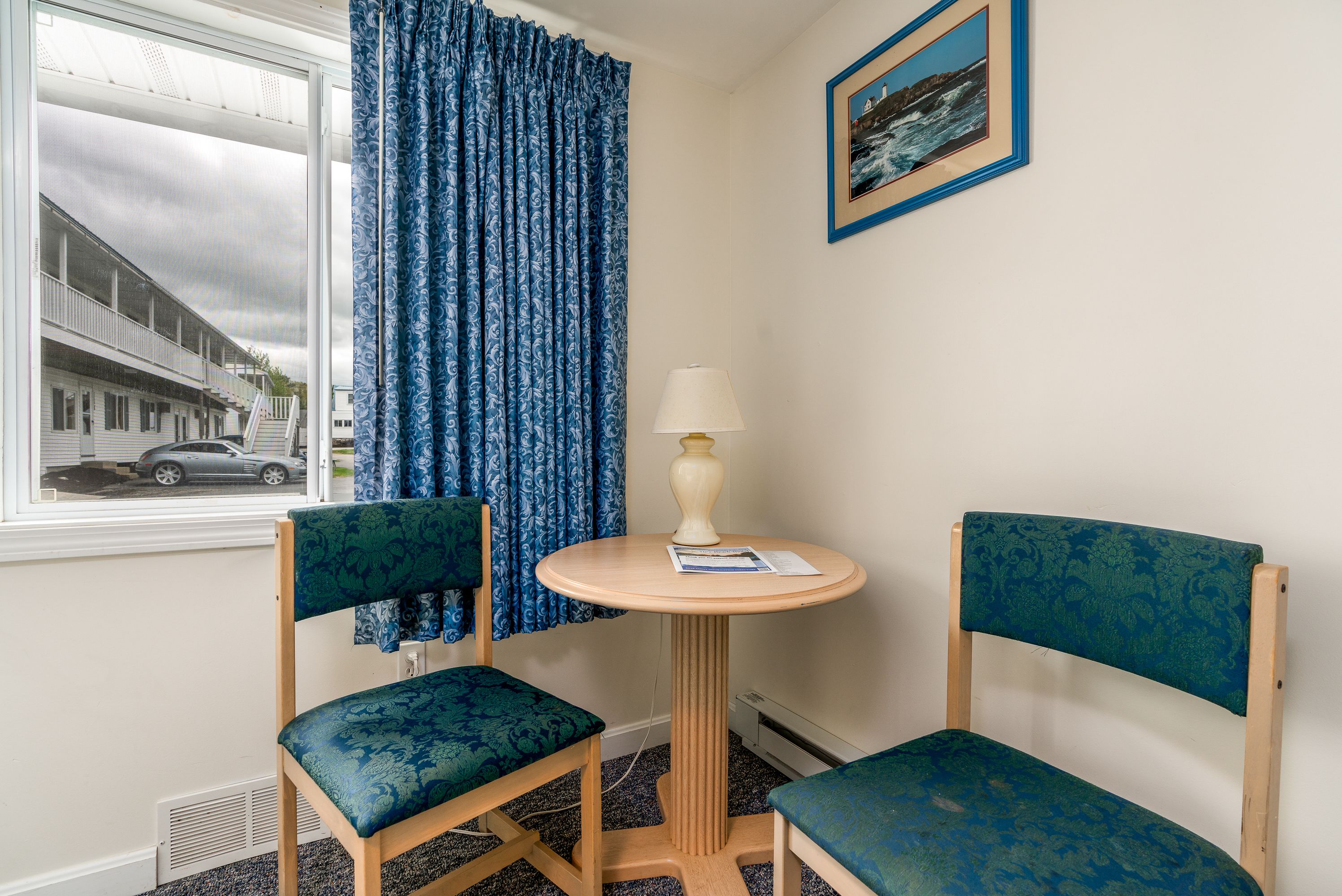 A corner room with two chairs, a small table with a lamp, a window with a view of a building, and a framed picture on the wall.