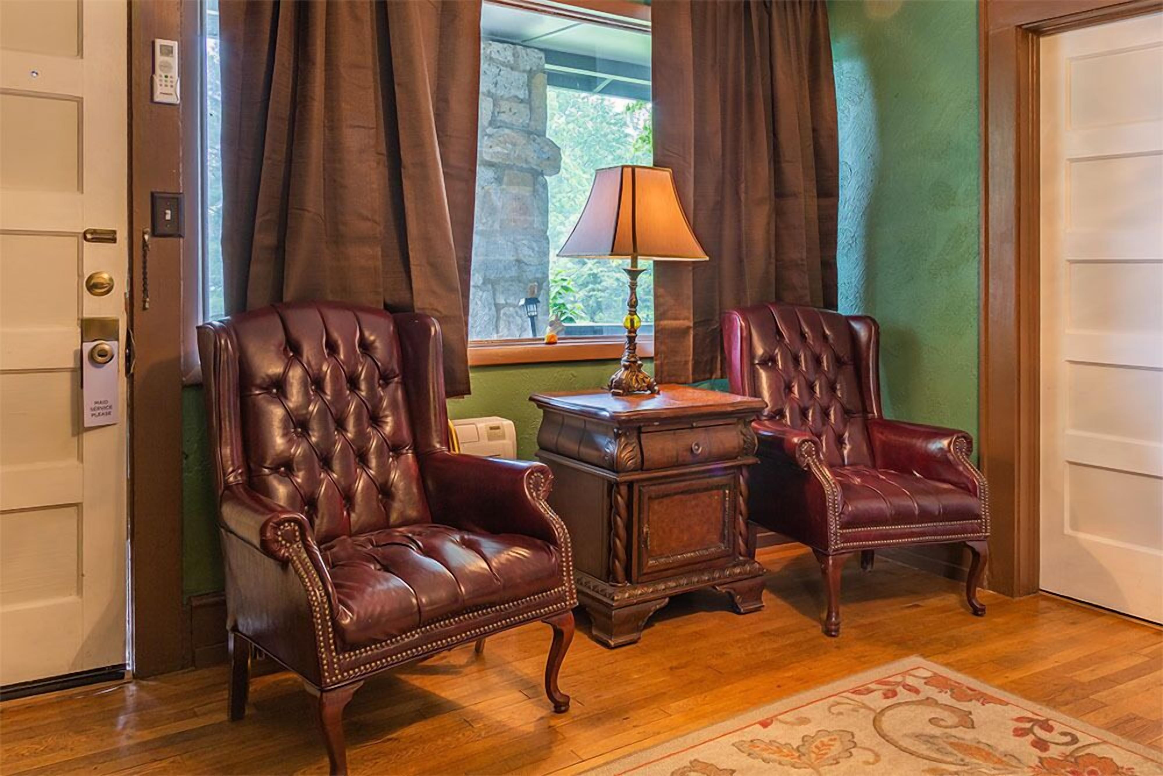 A room with two leather armchairs, a small wooden table with a lamp, and a view of greenery through a window.