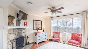 Une chambre avec une cheminée en pierre, un ventilateur de plafond, une grande fenêtre offrant une belle vue et deux fauteuils rouges.